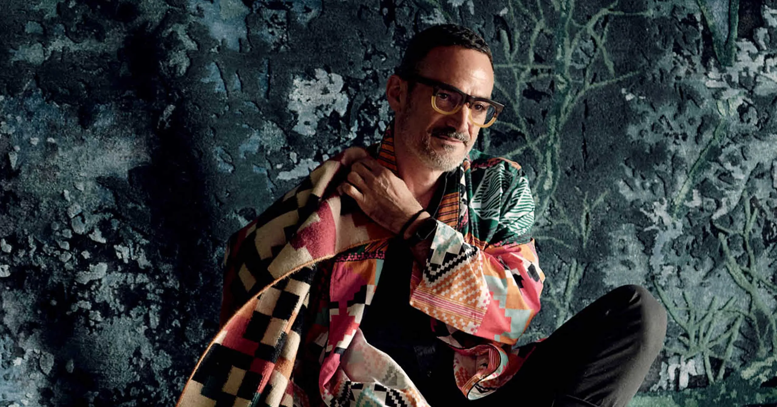 Man with short dark hair and glasses wearing a vibrant patchwork jacket, posing against a textured dark teal wall.