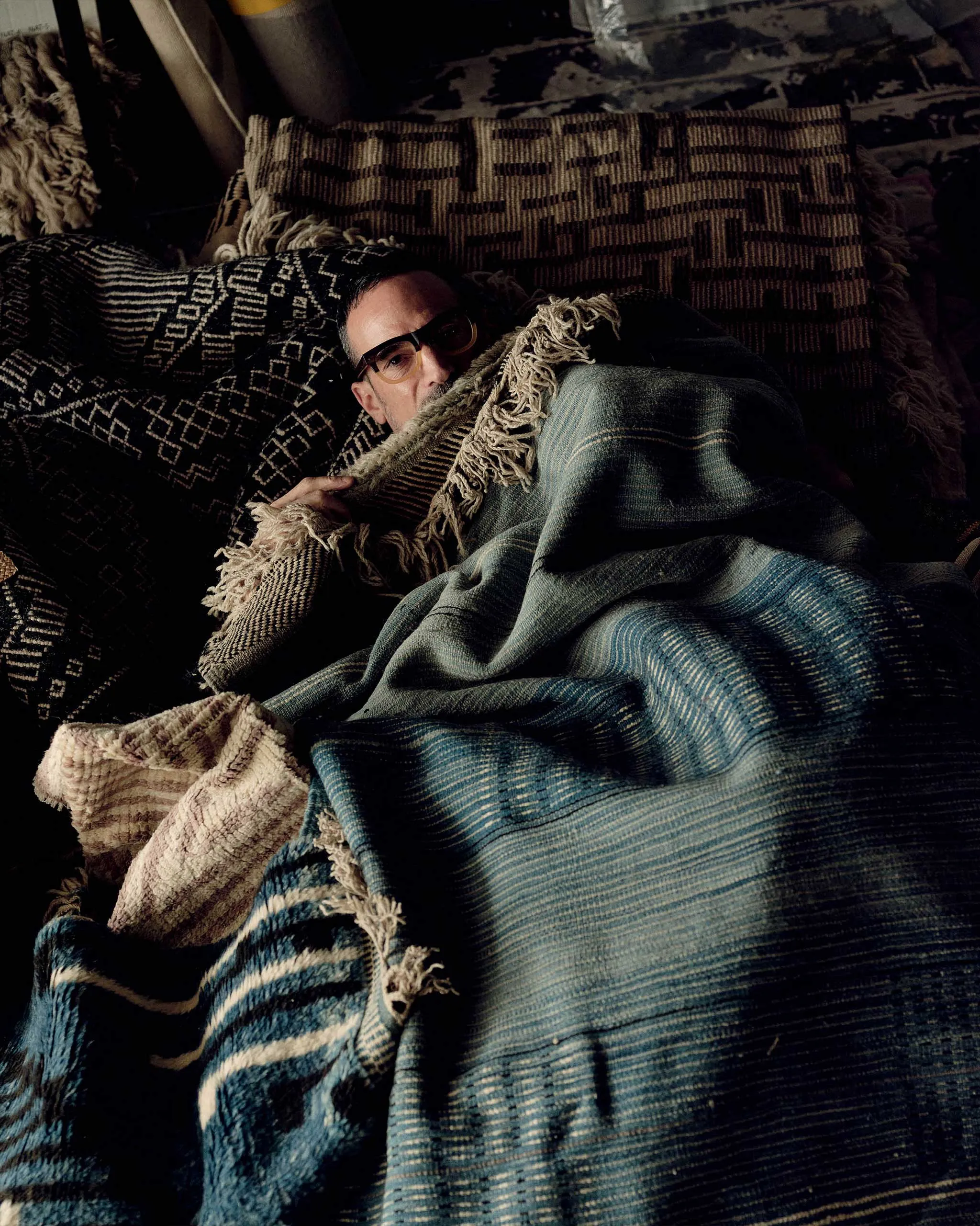 Person partially visible, lying under patterned blankets, wearing glasses in a dimly lit room.