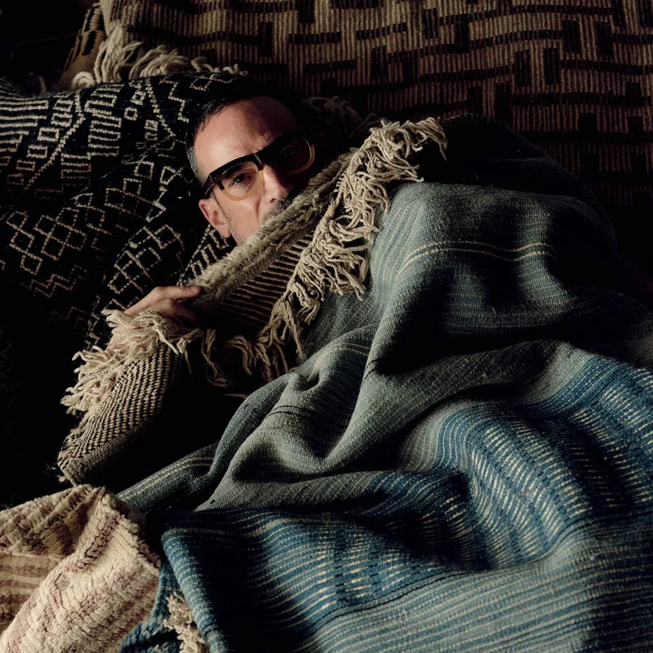 Man with short dark hair and glasses wearing a vibrant patchwork jacket, posing against a textured dark teal wall.