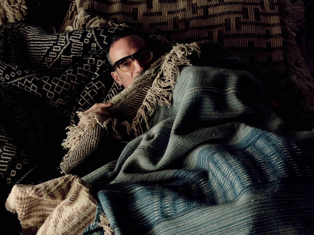 Man with short dark hair and glasses wearing a vibrant patchwork jacket, posing against a textured dark teal wall.