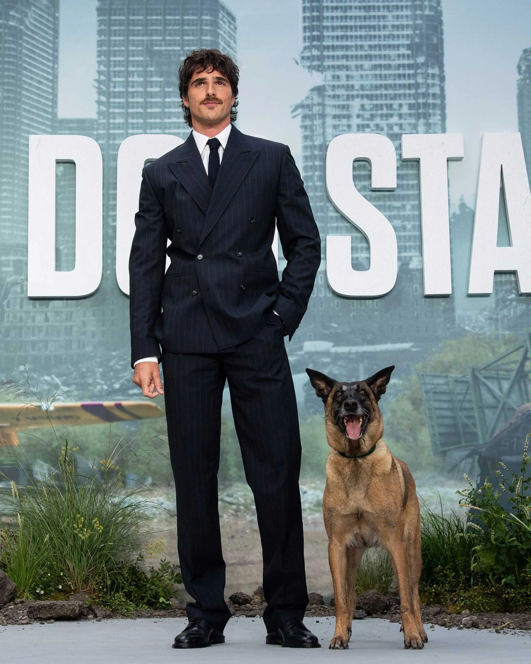 Man in a dark pinstripe suit posing with a German Shepherd dog outdoors, with large white letters and city buildings in the background.