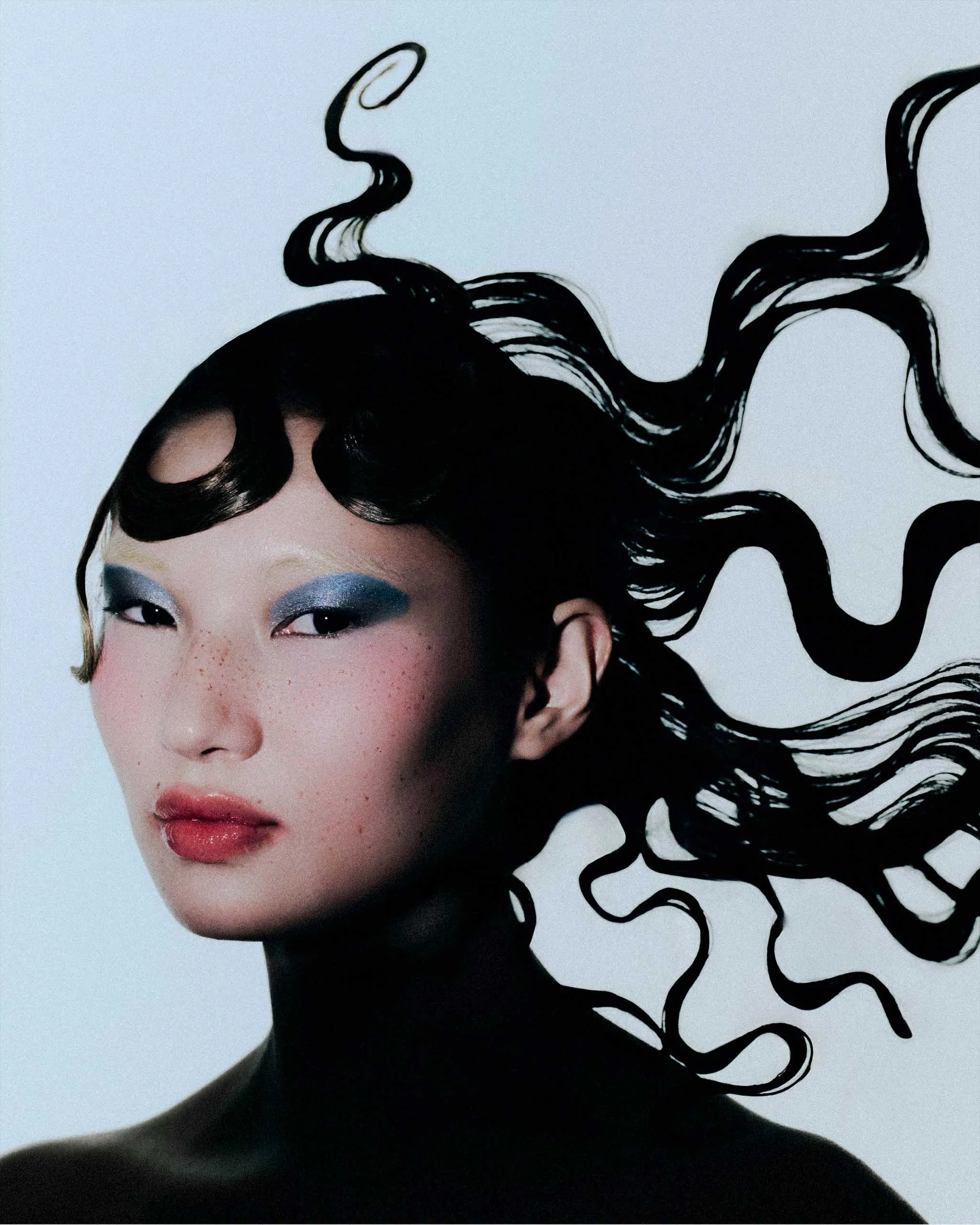 Close-up portrait of a person with blue eye makeup, freckles, pink lips, and flowing black hair against a pale blue background.