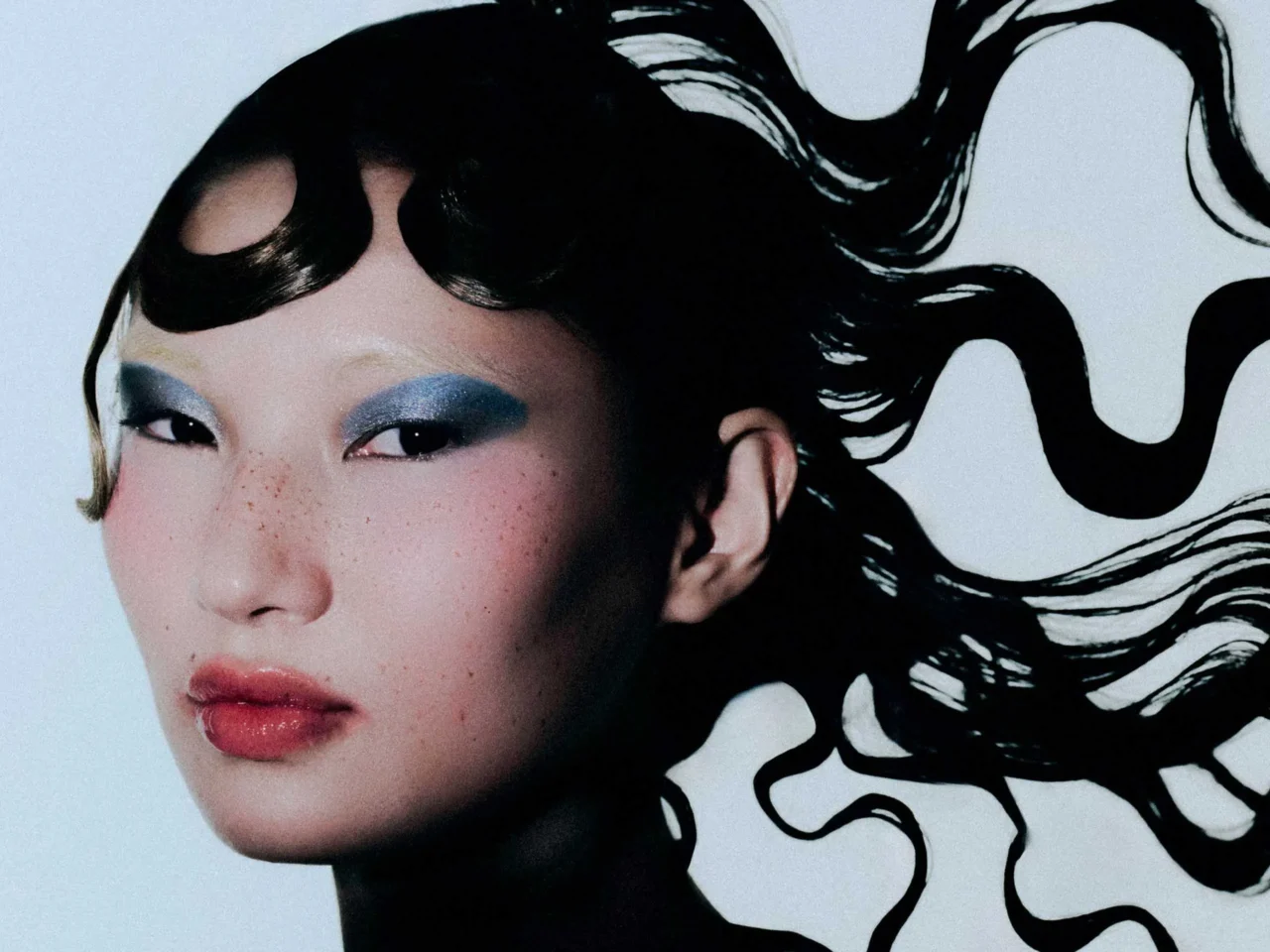 Close-up portrait of a woman with blue eyeshadow, freckles, pink lips, and flowing black hair against a pale background.