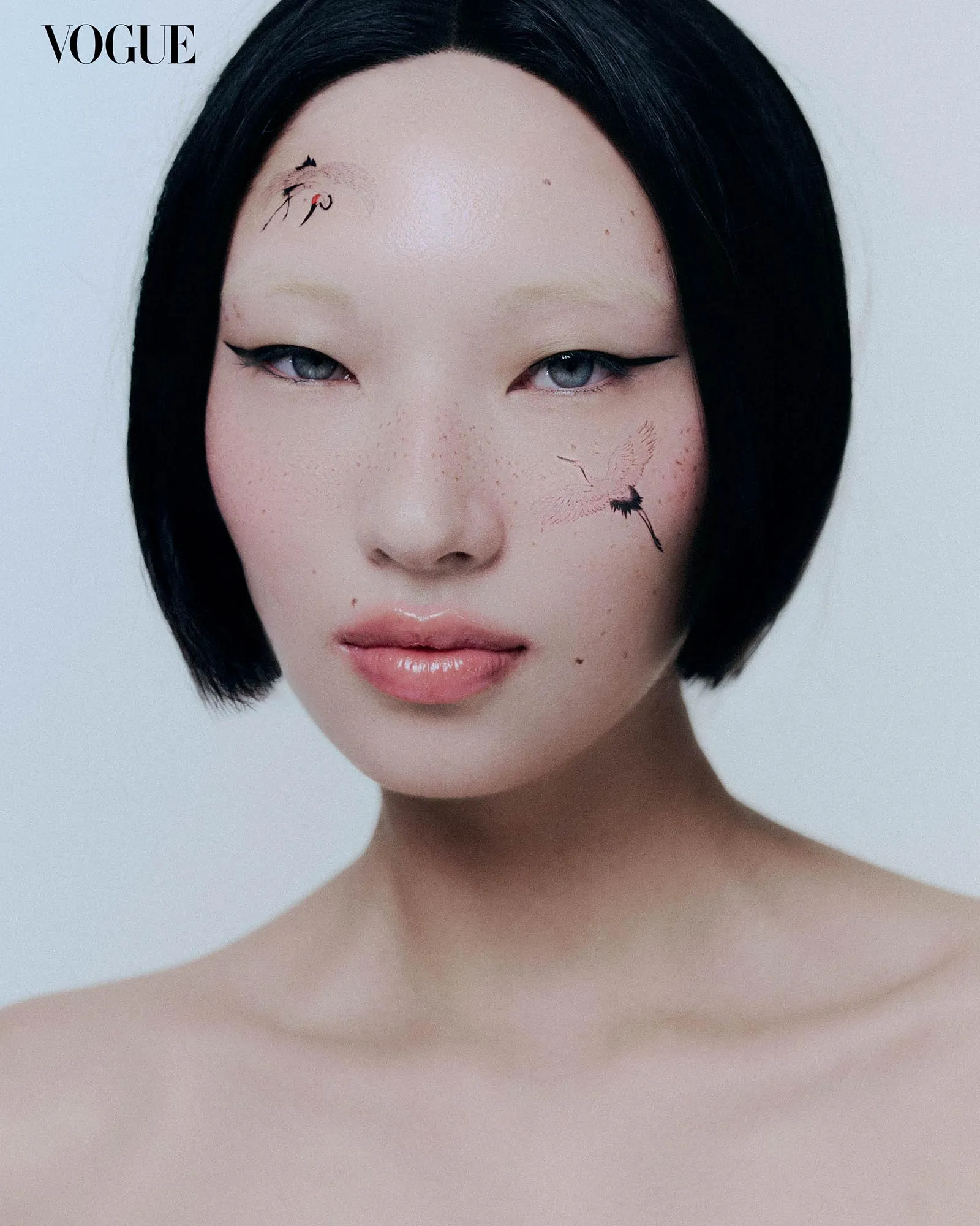Close-up portrait of a woman with short black hair, pale skin, blue eyes, winged eyeliner, and dandelion tattoos on her temple and cheek; Vogue logo in the corner.