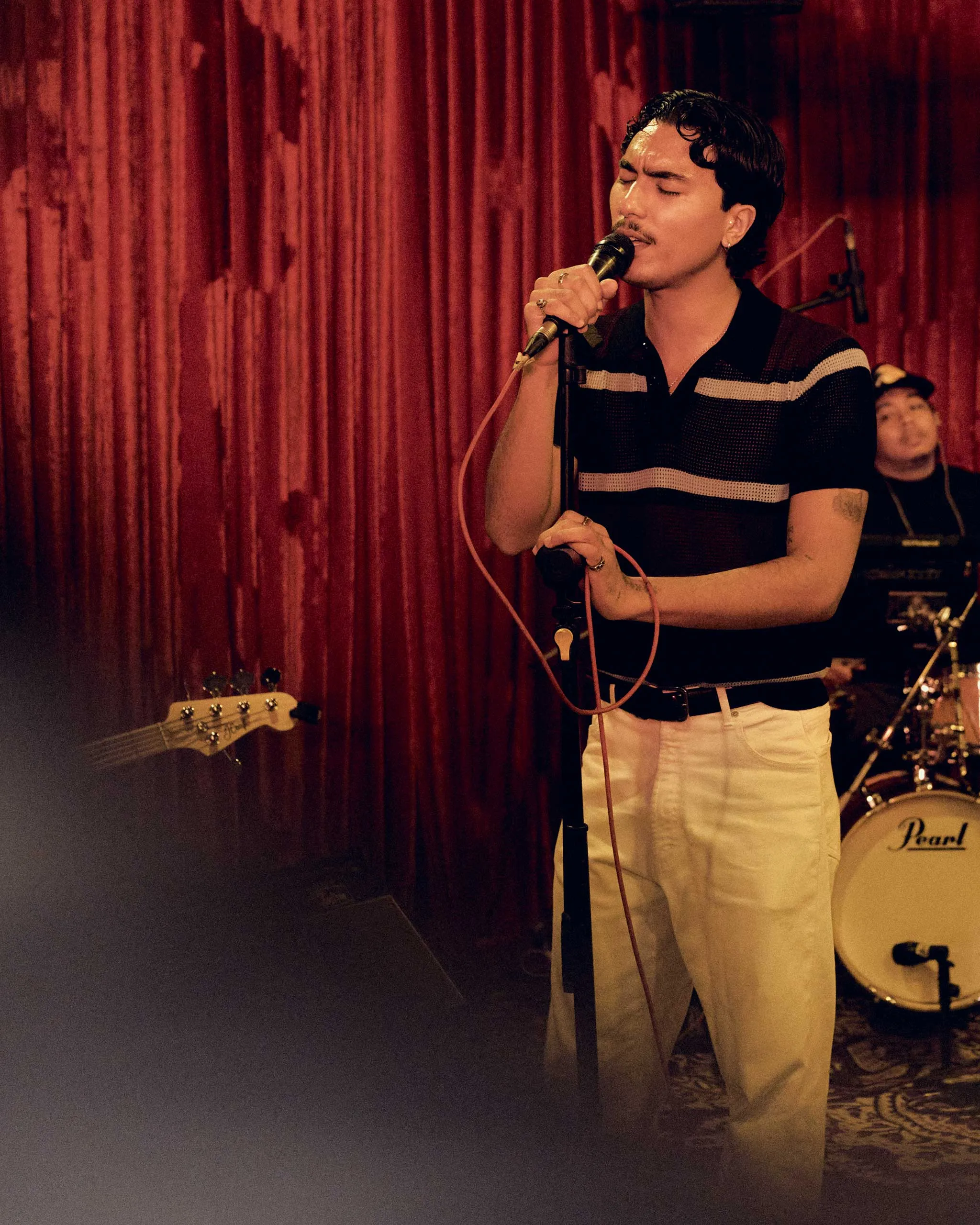 Male singer on stage gripping a microphone, eyes closed, performing in front of red curtains with a guitar visible in the foreground and drums in the back.