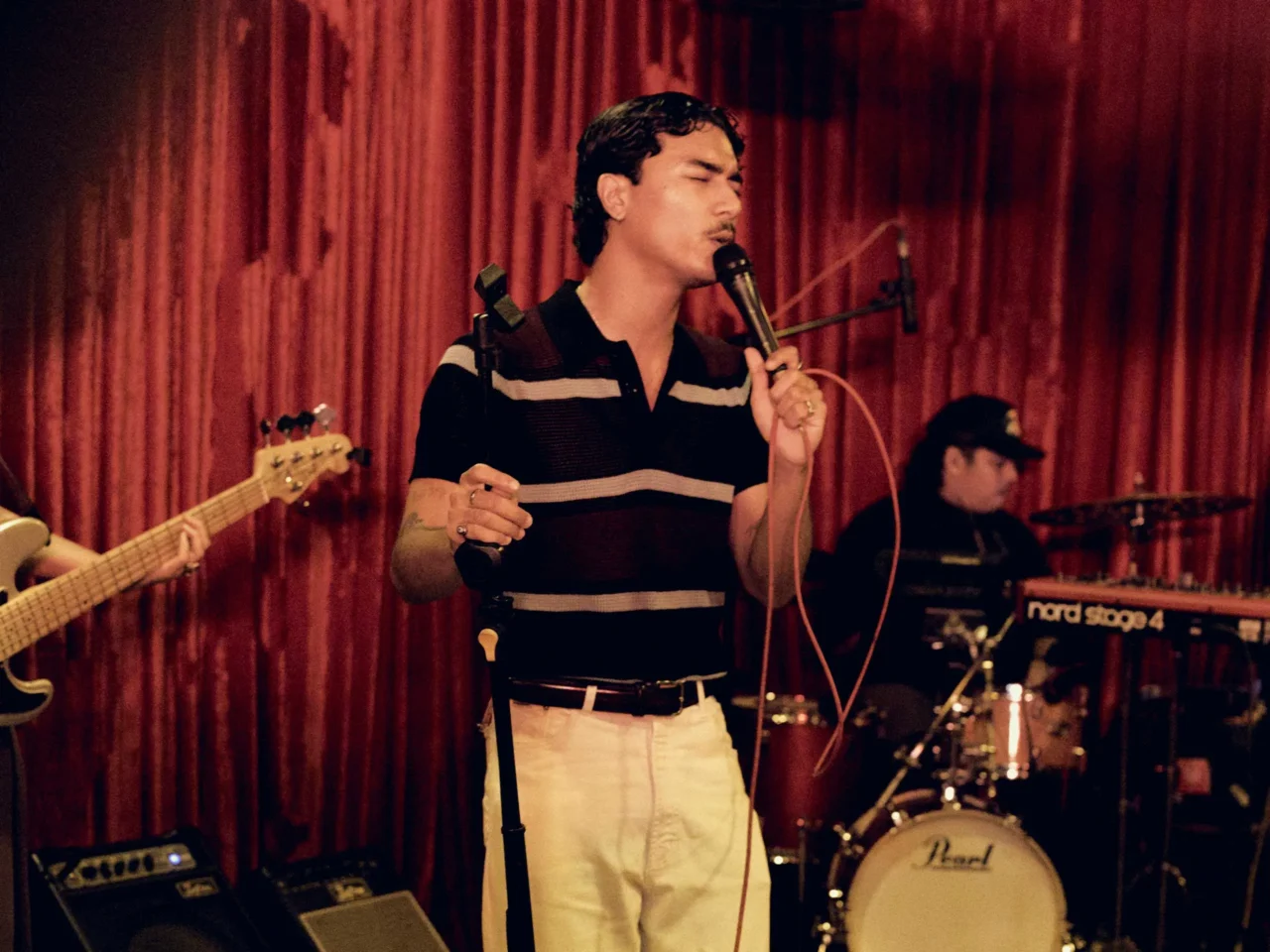 Male singer on stage with eyes closed, mic in hand, red curtain backdrop, mid-performance