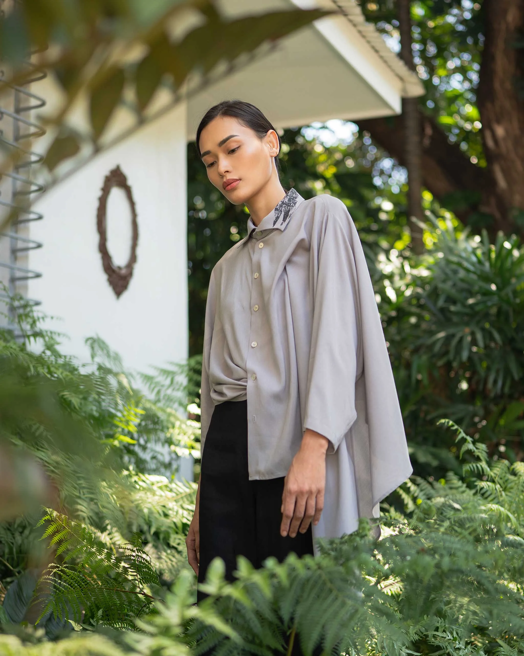 Woman standing in a lush garden wearing an oversized gray shirt with a patterned collar, looking down thoughtfully amid green ferns and foliage.