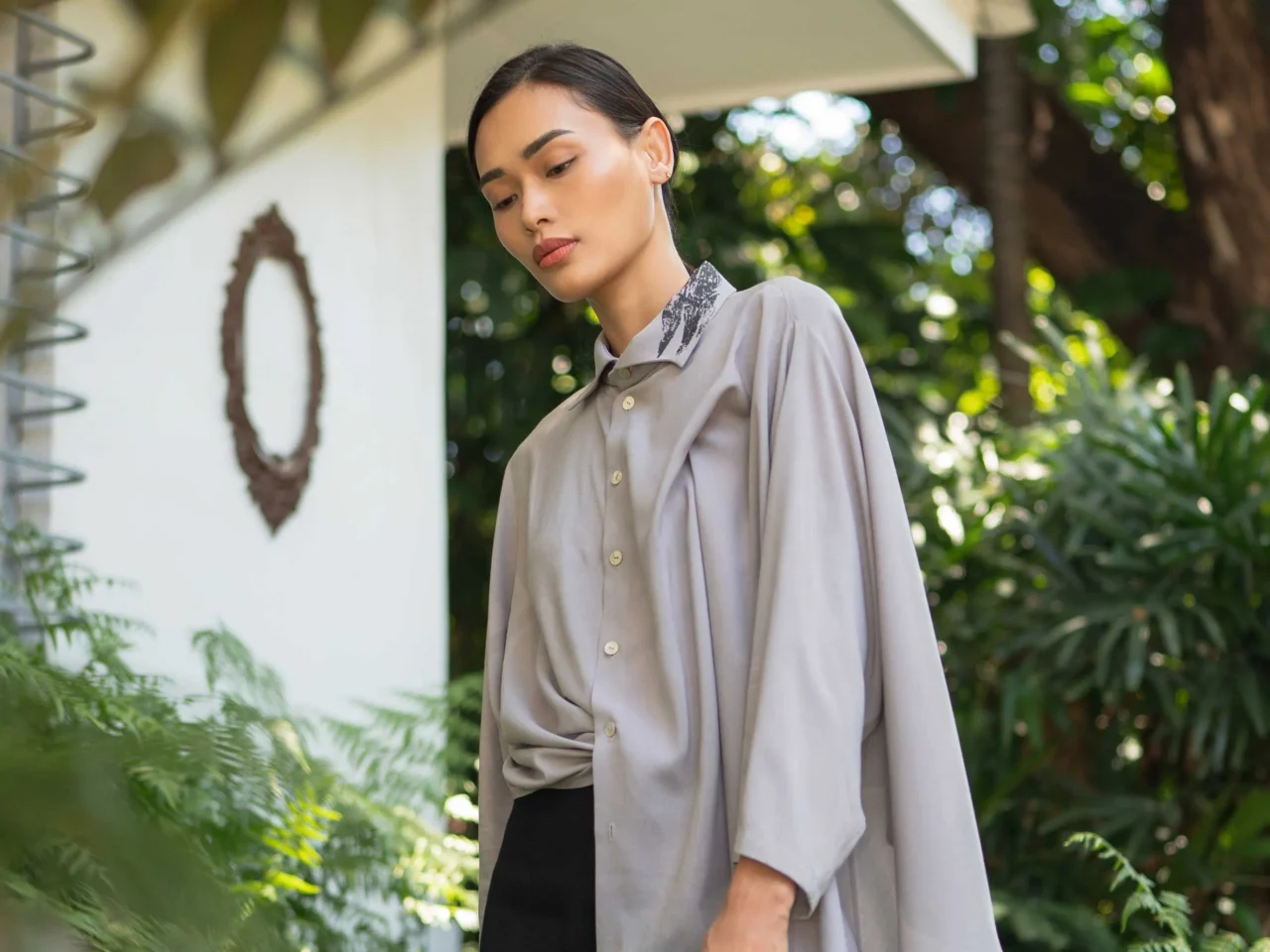 Woman in a white blouse and a patterned gray-black skirt walks along a balcony with green plants in the background.
