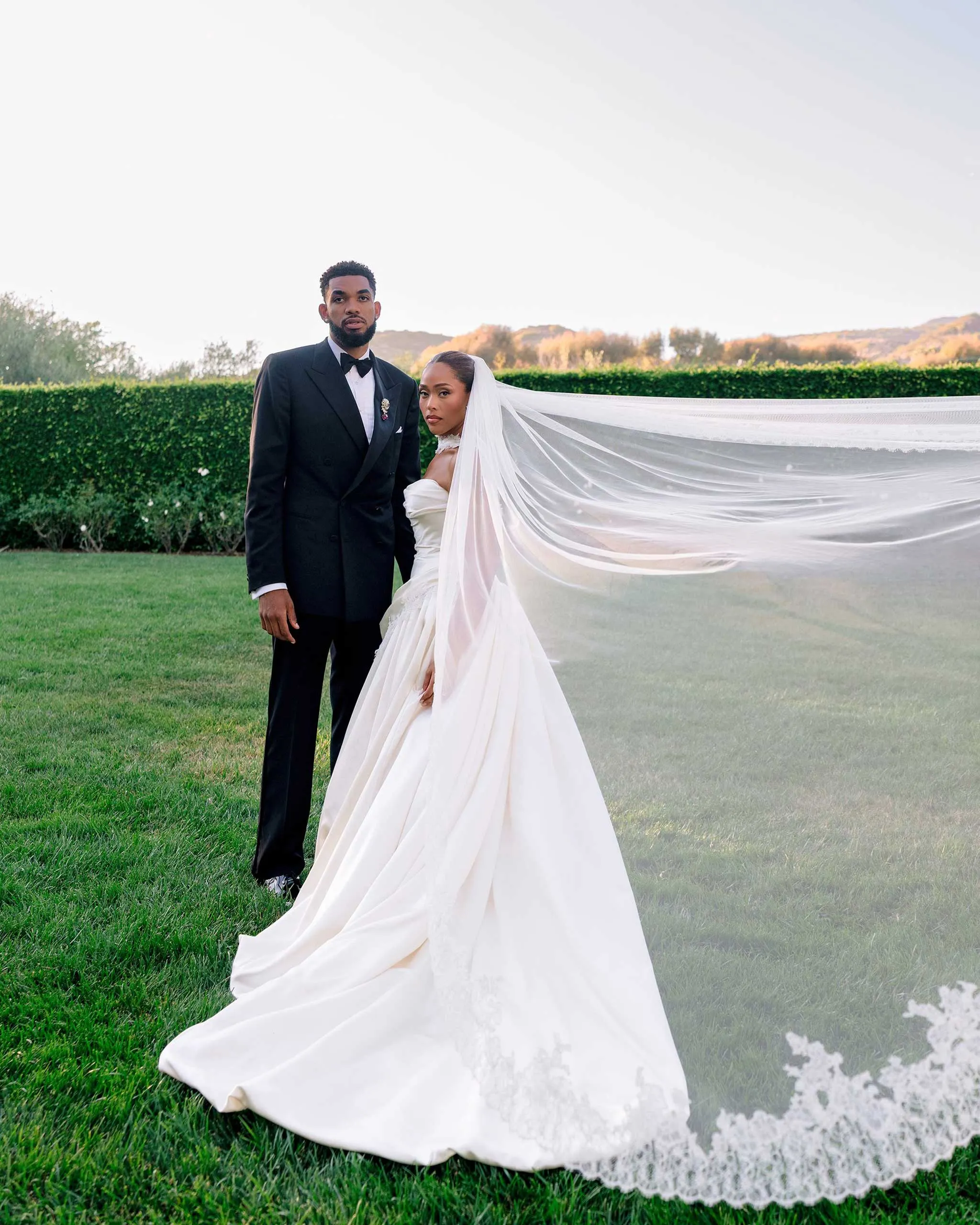 A bride in a white wedding dress with a long veil stands beside a man in a black tuxedo on a grassy lawn.