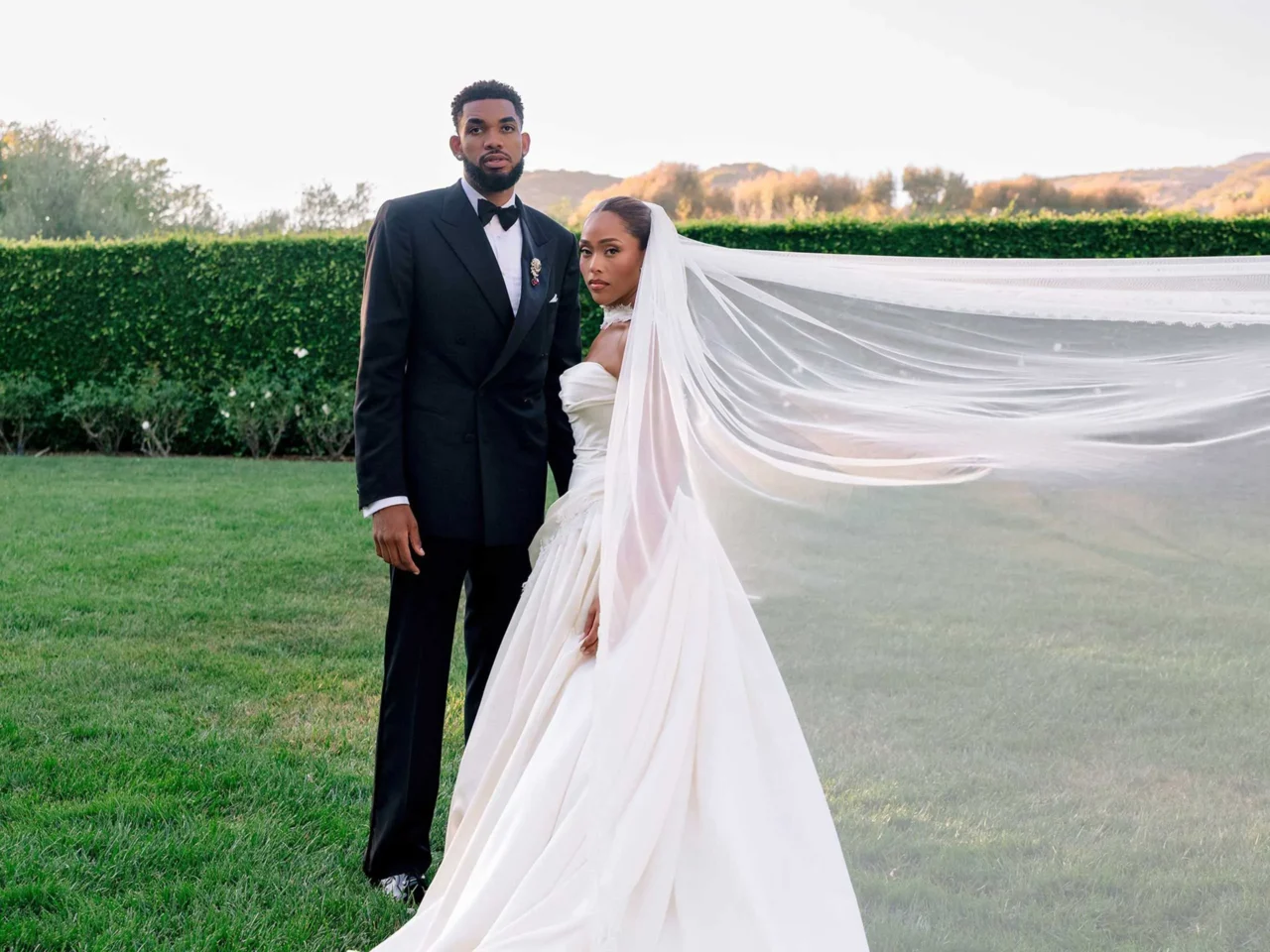 Bride in a white wedding gown with a long veil stands beside a groom in a black tuxedo on a grassy lawn, hedge and hills in the background.