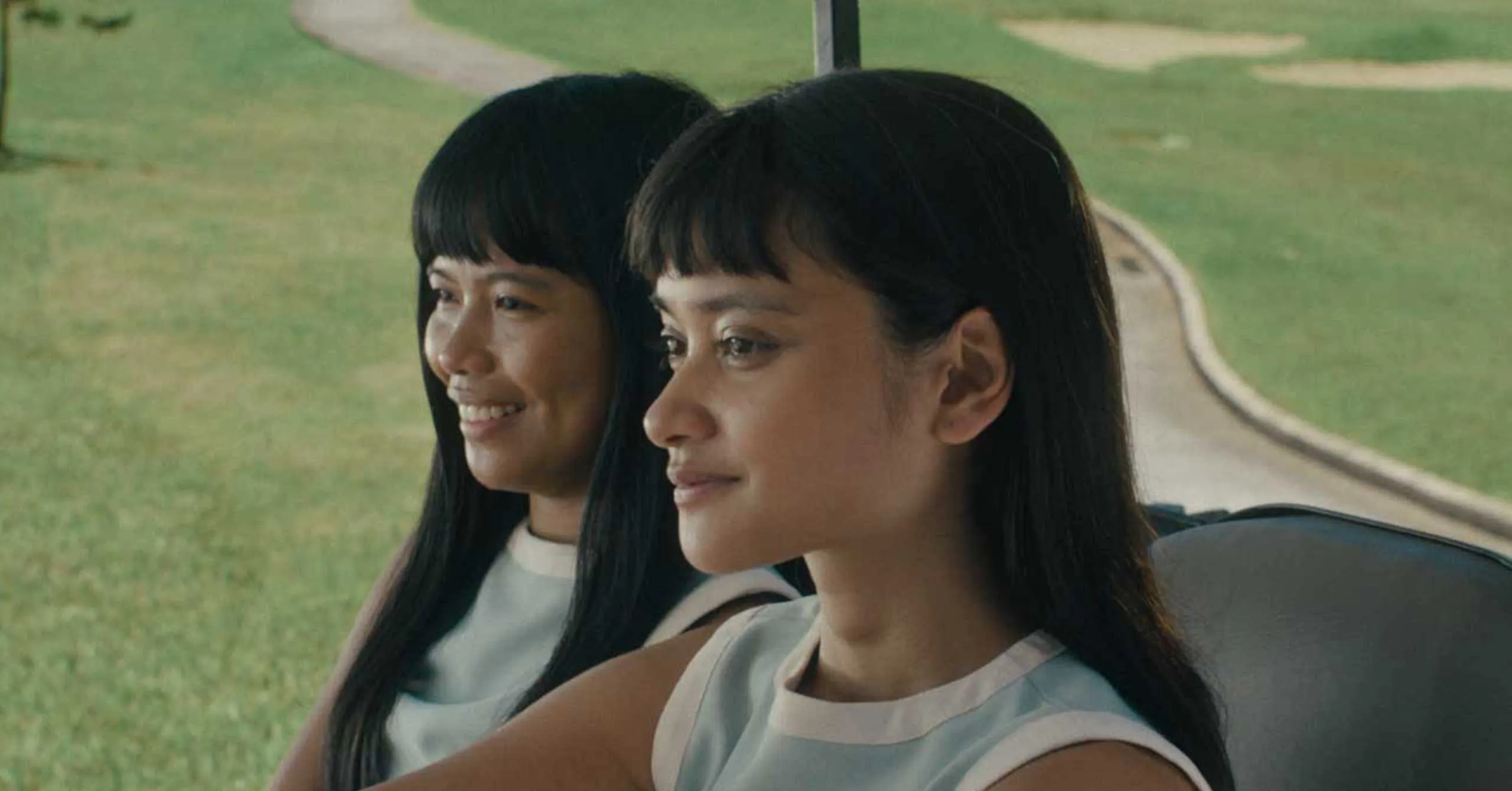 Two women with dark hair sitting in a car, side by side, smiling, with a grassy park outside the window.