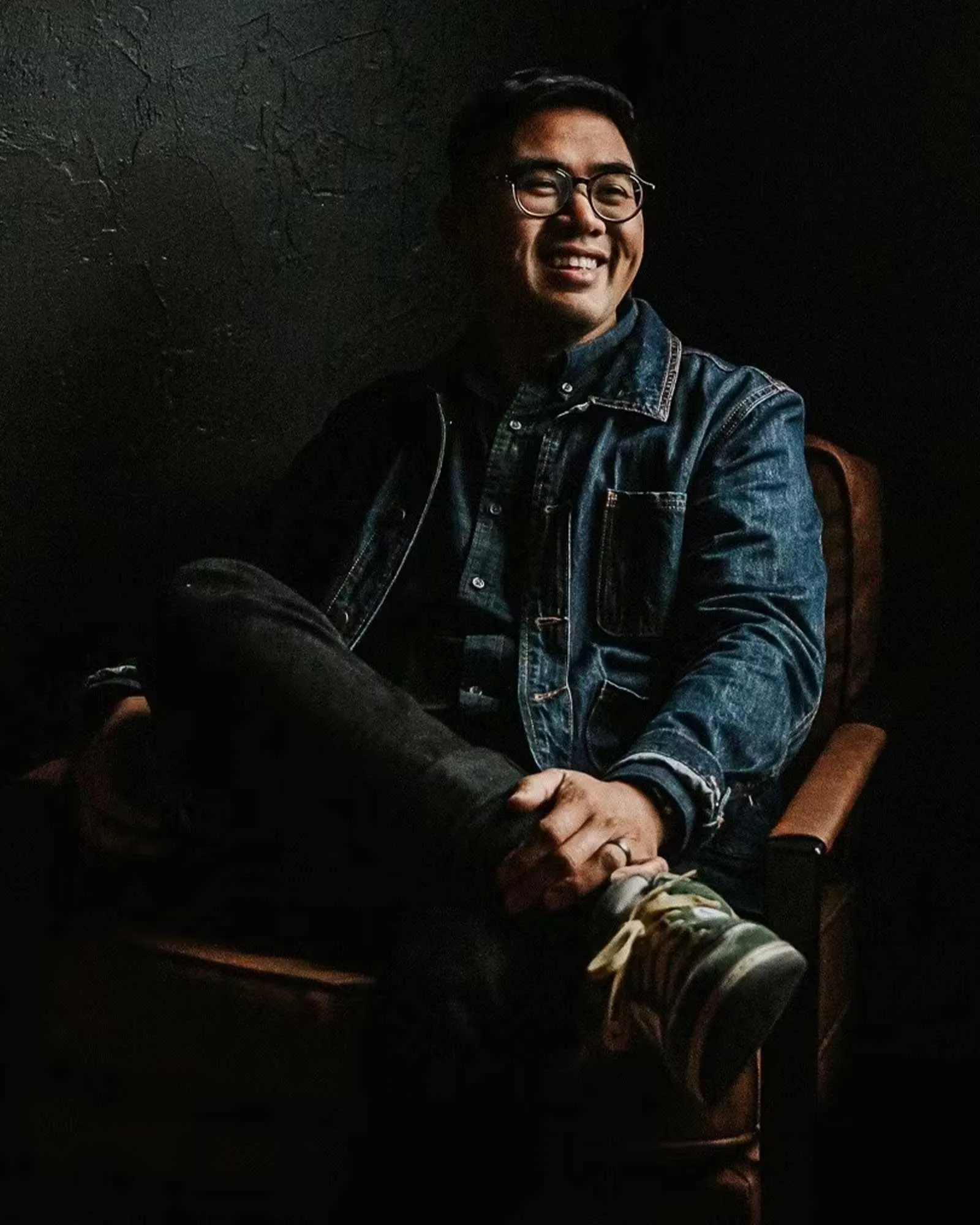 Smiling man wearing glasses and a denim jacket sits in a dark room with a textured wall behind him.