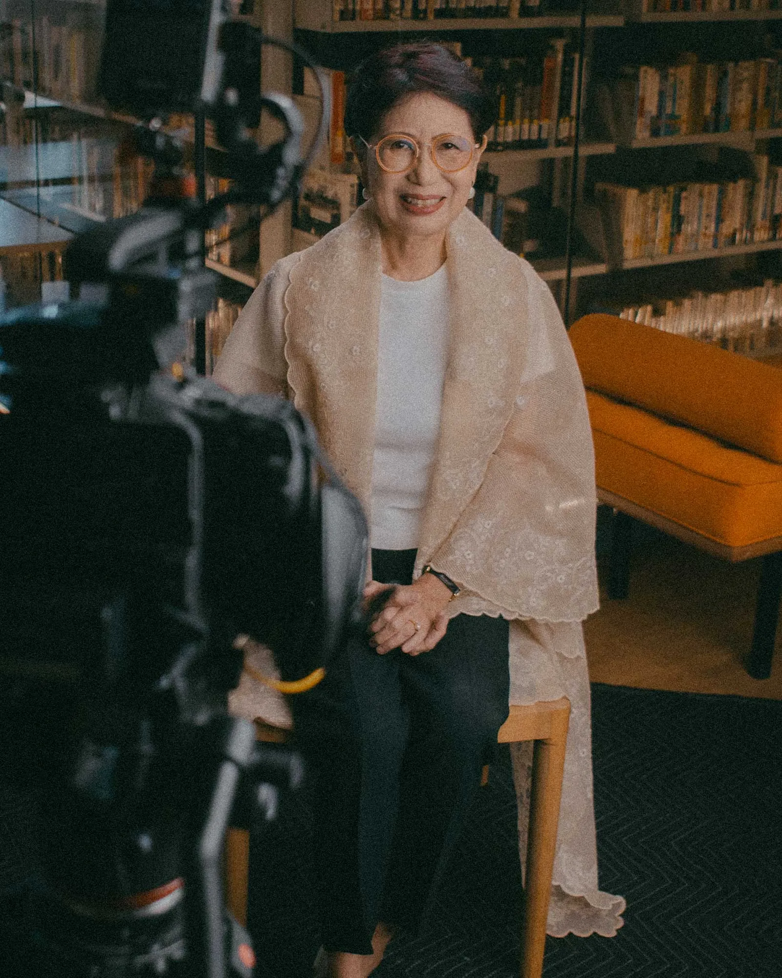 Smiling older woman with round glasses sits in a library-like setting, wearing a light lace shawl over a white top while a camera looms in the foreground for a video shoot.