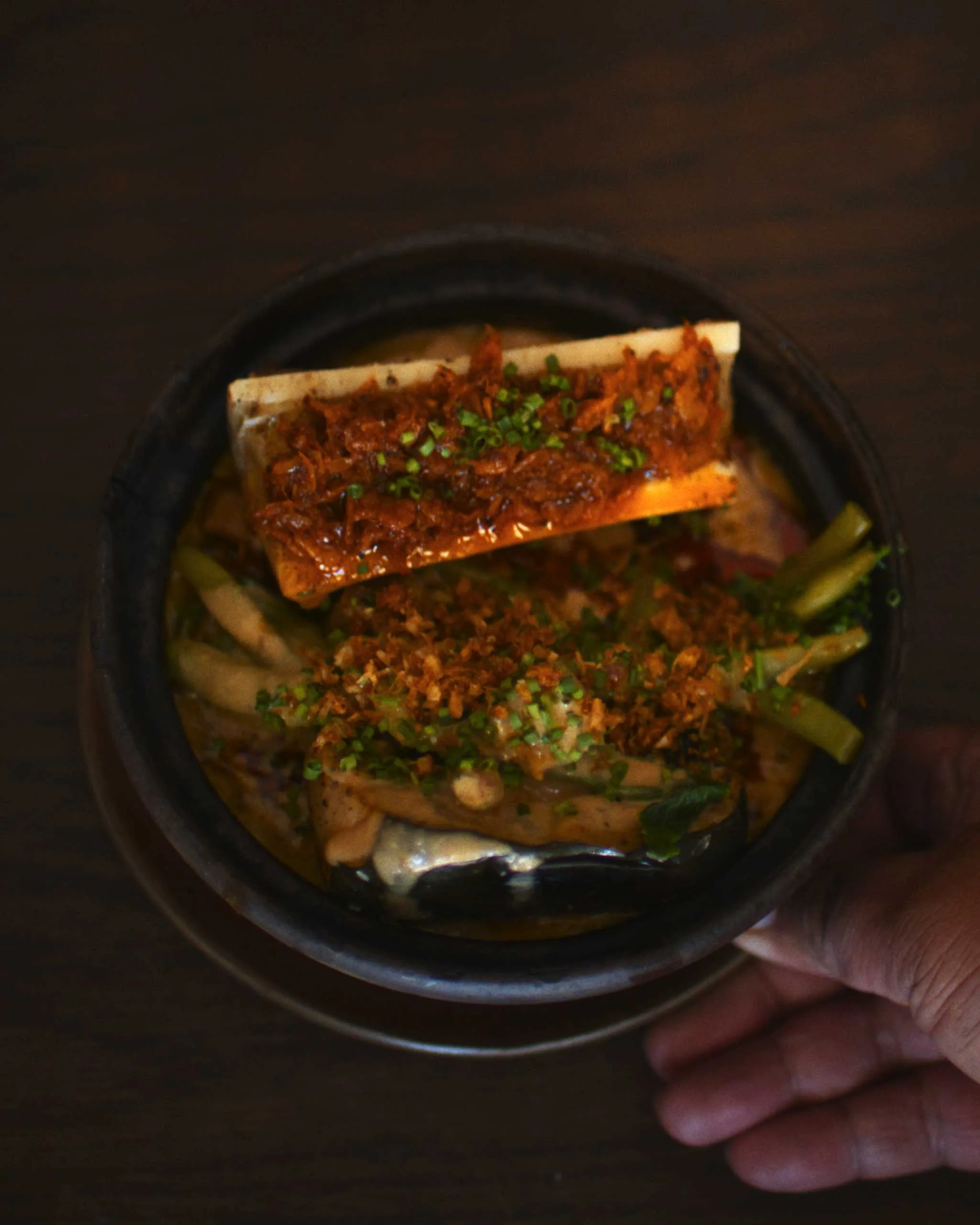 Close-up of a dark bowl with a fried, saucy topping on a rectangular piece of food, garnished with chopped green onions, held in a hand.
