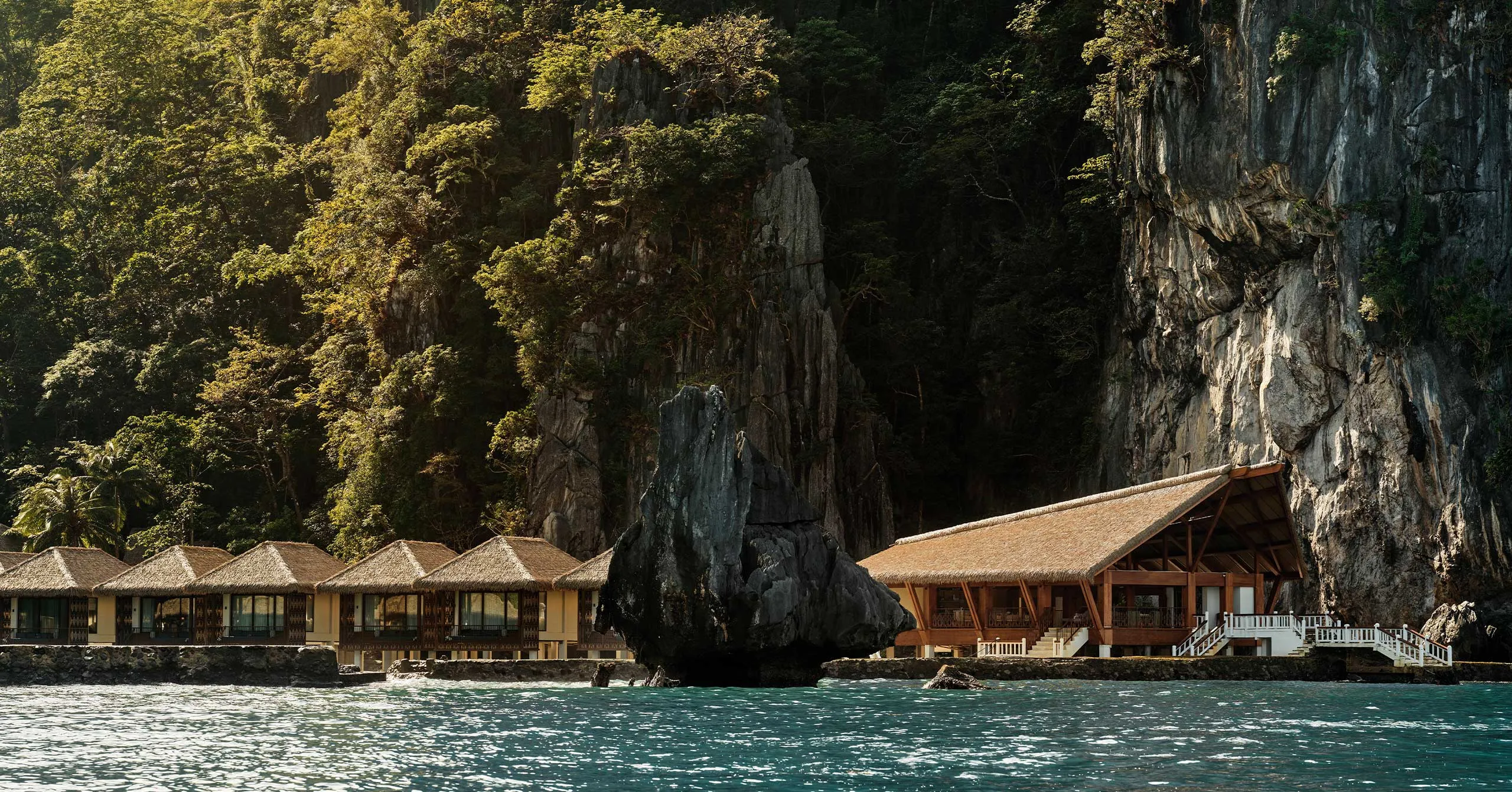 Resort buildings with thatched roofs along a rocky cliff by clear blue water.