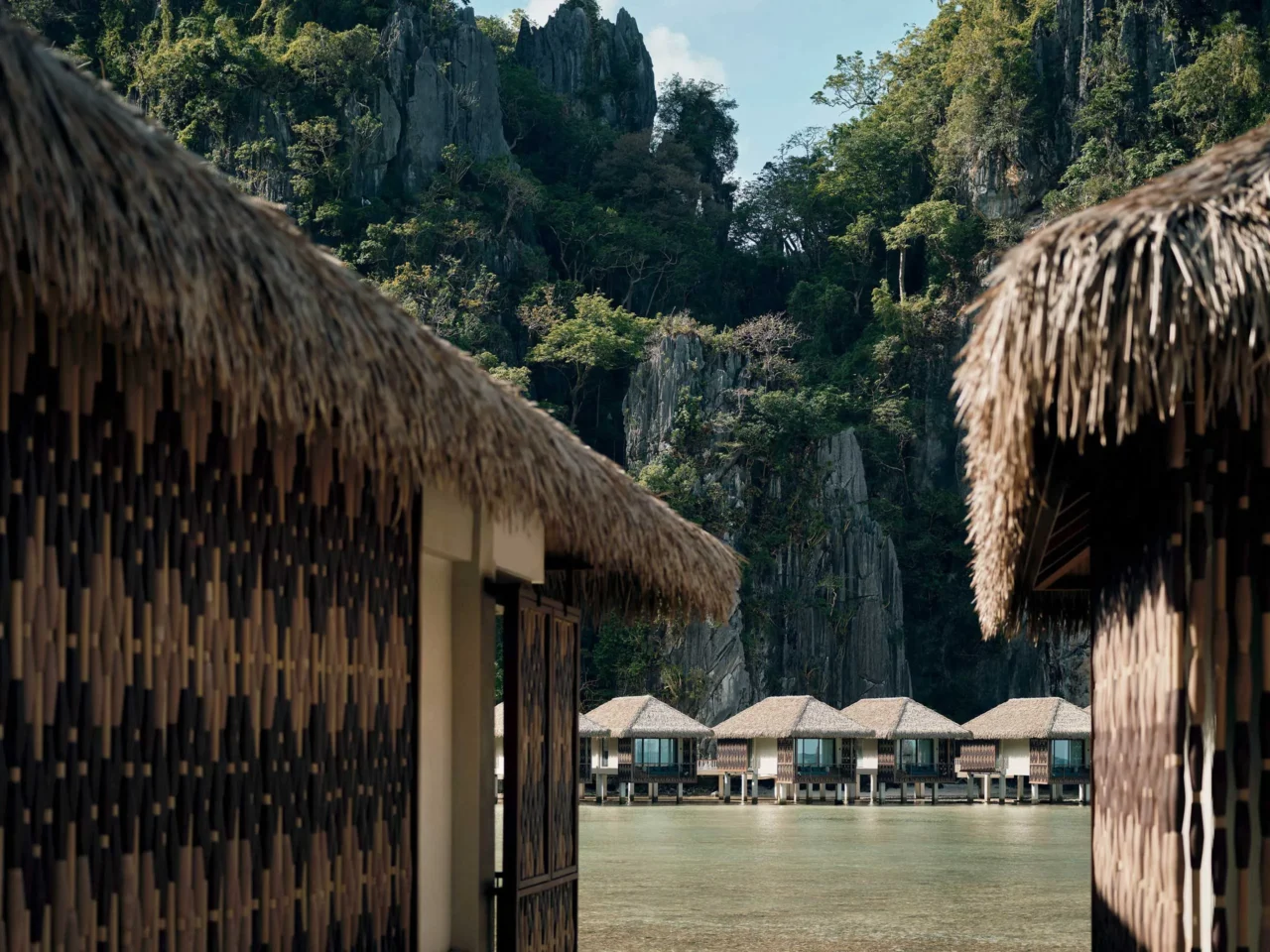 Resort buildings with thatched roofs along a rocky cliff by clear blue water.