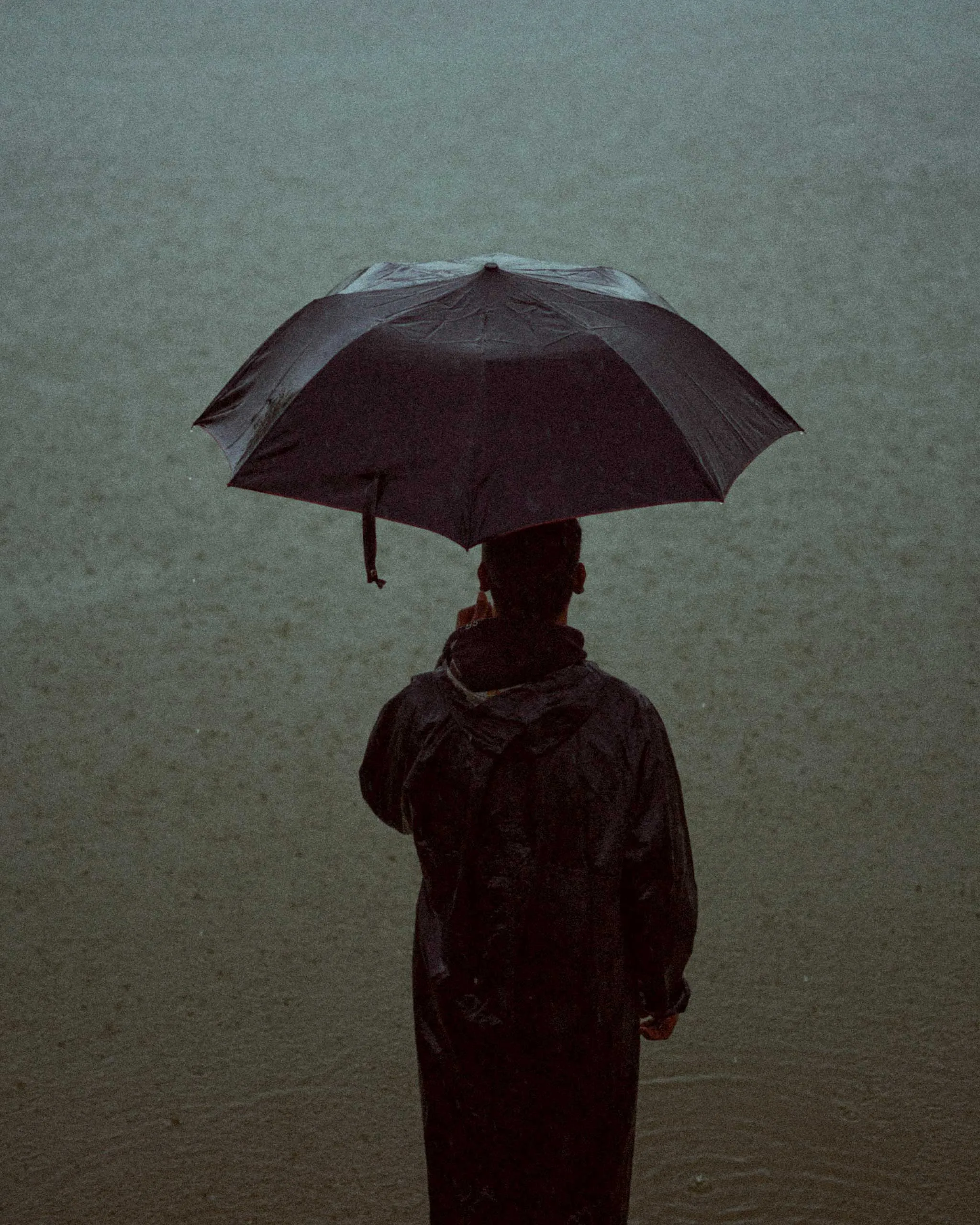Back view of a person wearing a rain jacket on a sandy beach, holding a large black umbrella in a rainy, overcast scene.