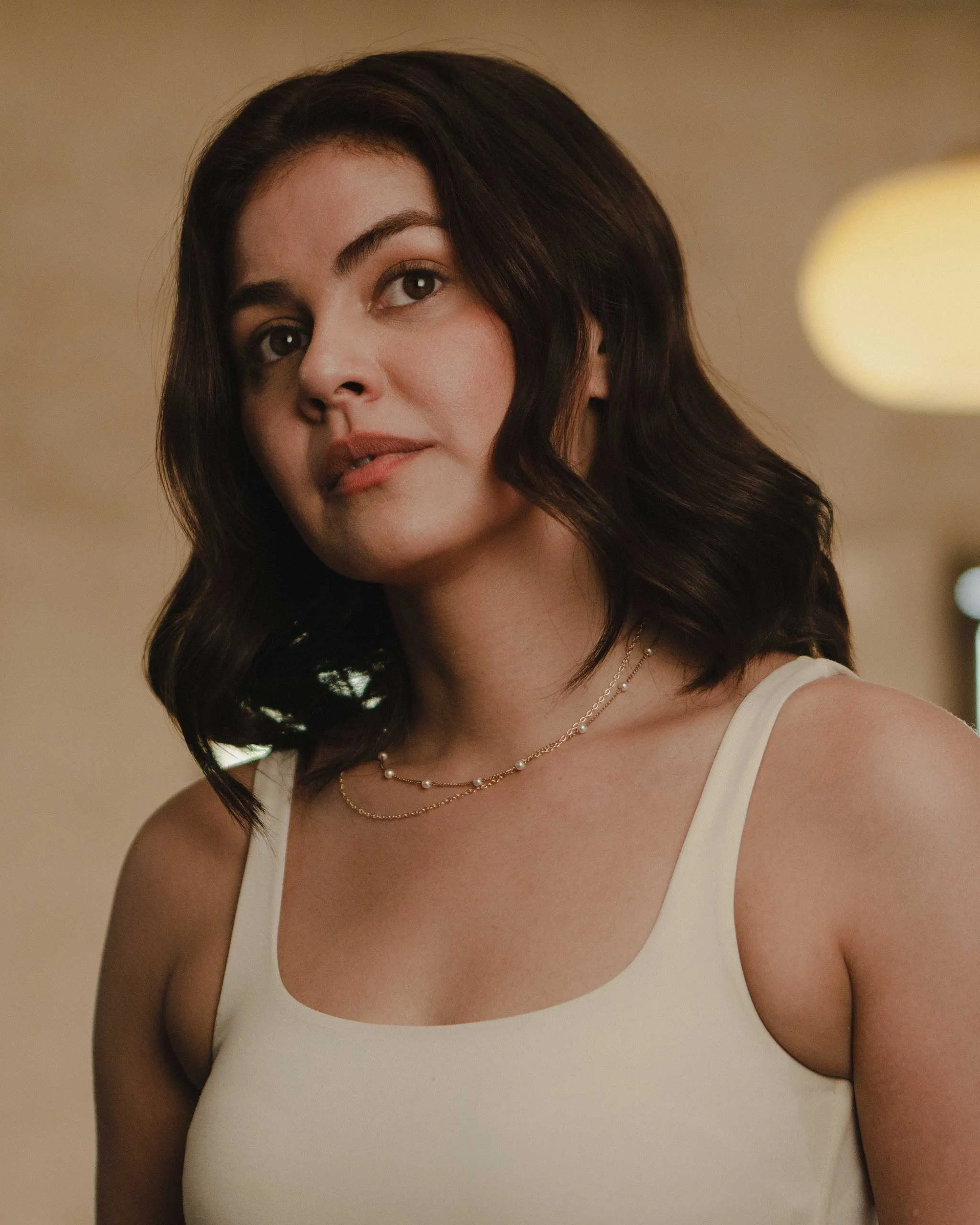 Close-up portrait of a woman with dark hair, wearing a white sleeveless top and a delicate pearl necklace in warm indoor lighting.