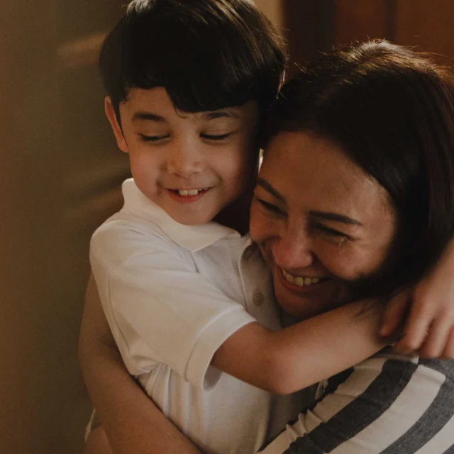A child and adult share a warm hug, both smiling with their eyes closed in a close, affectionate moment.