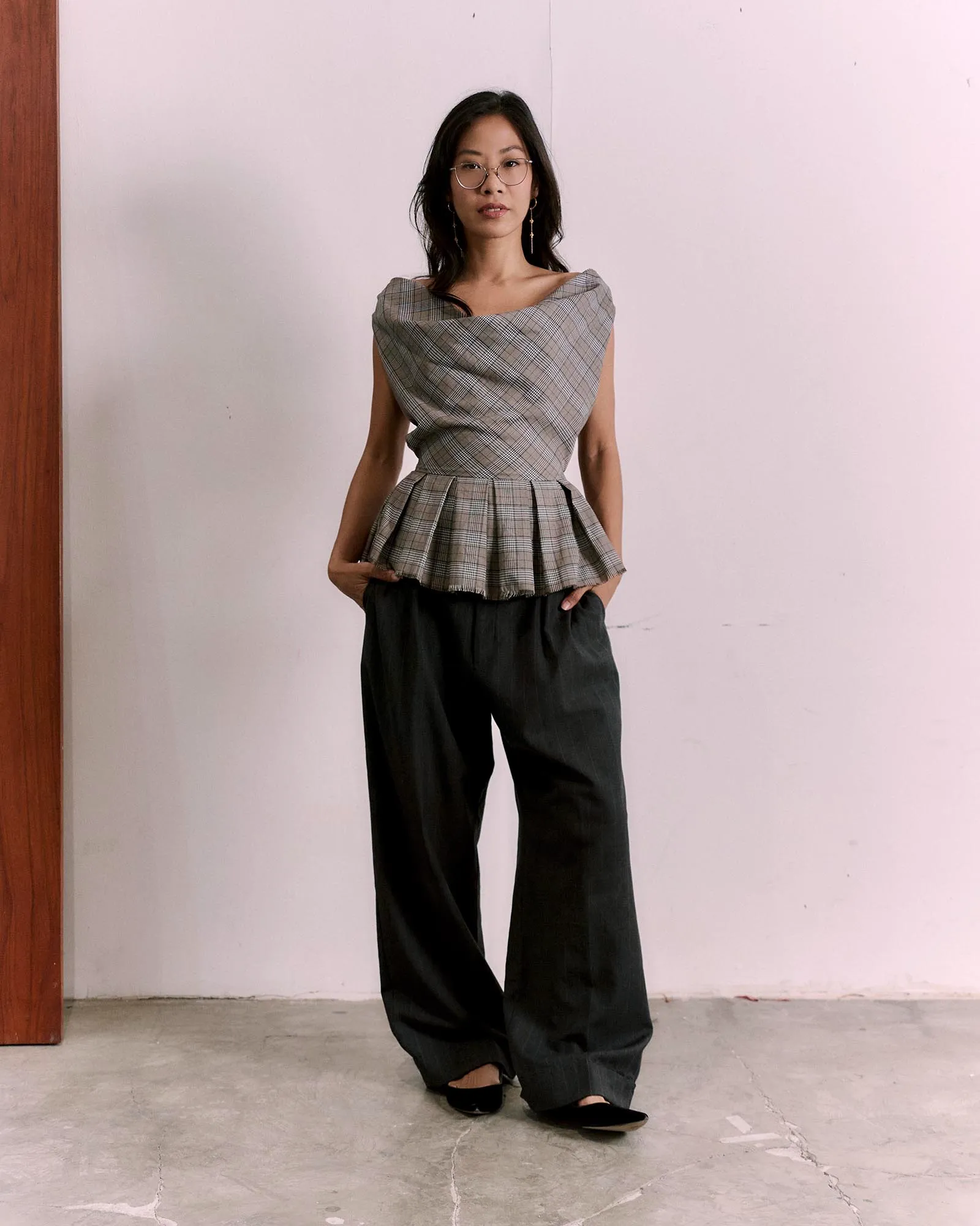 Woman standing indoors against a pale wall, wearing a gray plaid off-shoulder top with a layered peplum and black wide-leg pants, hands in pockets.