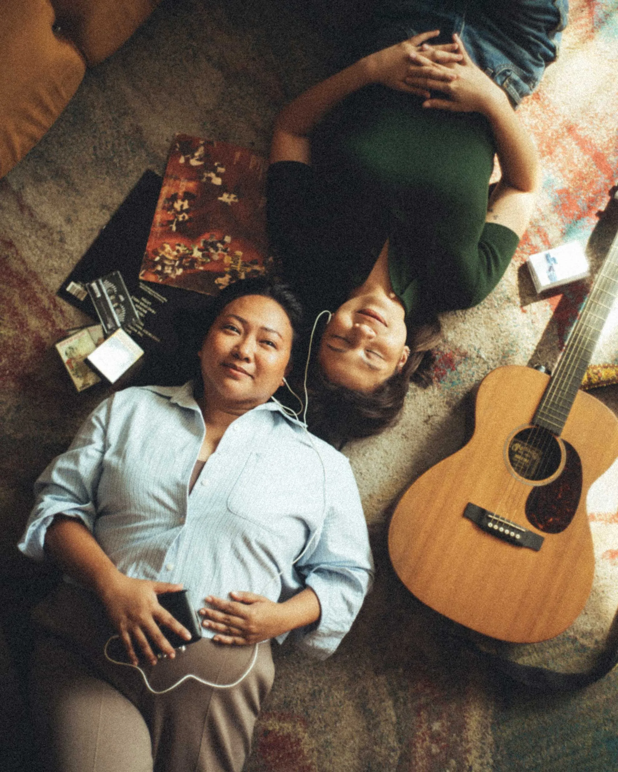 Two friends lying on a colorful rug with an acoustic guitar beside them.
