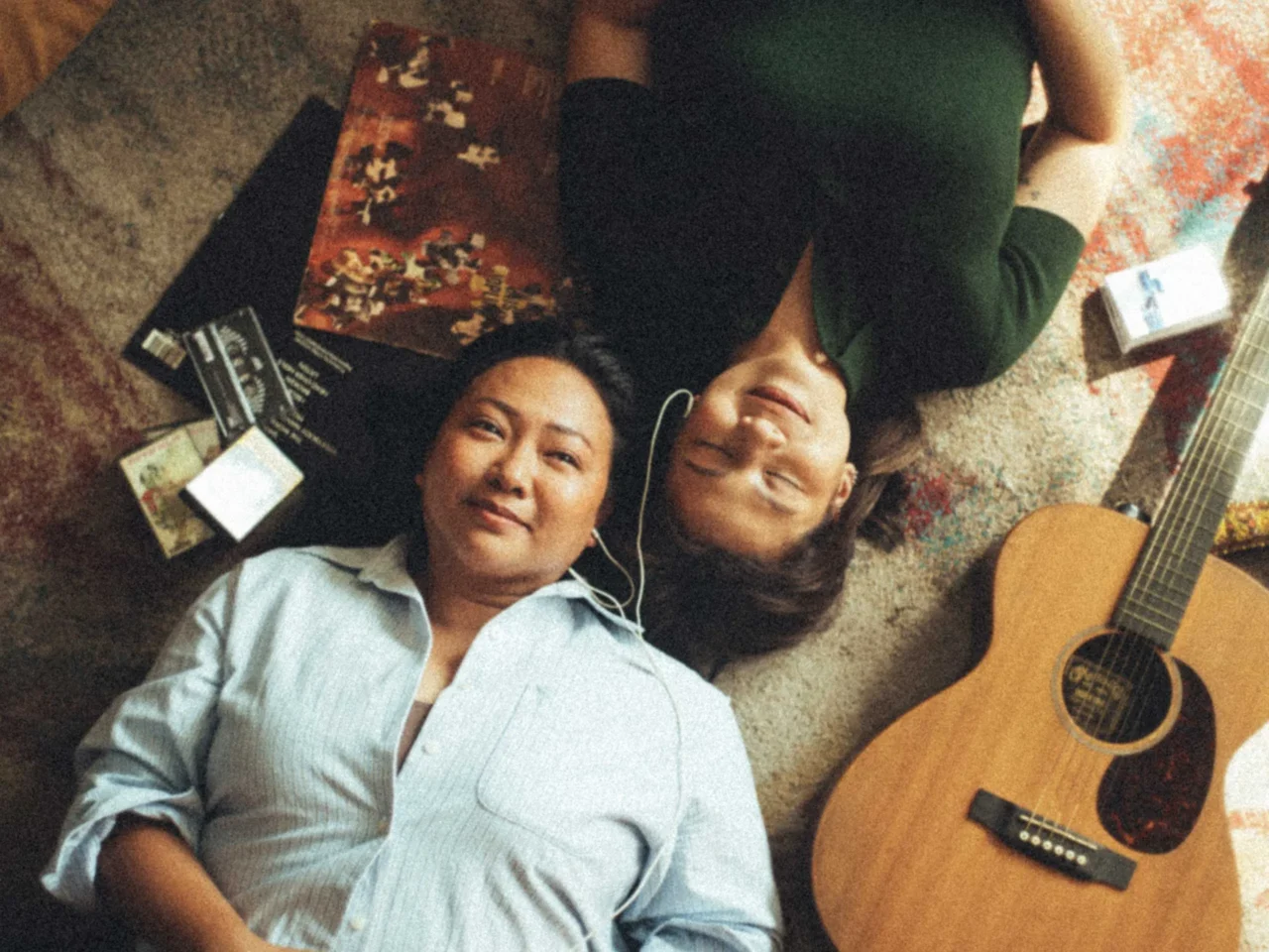 Two people lying on a colorful rug, smiling at the camera with a guitar nearby and earbud cables visible.