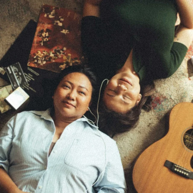 Two people lying on a colorful rug, smiling, with an acoustic guitar beside them.