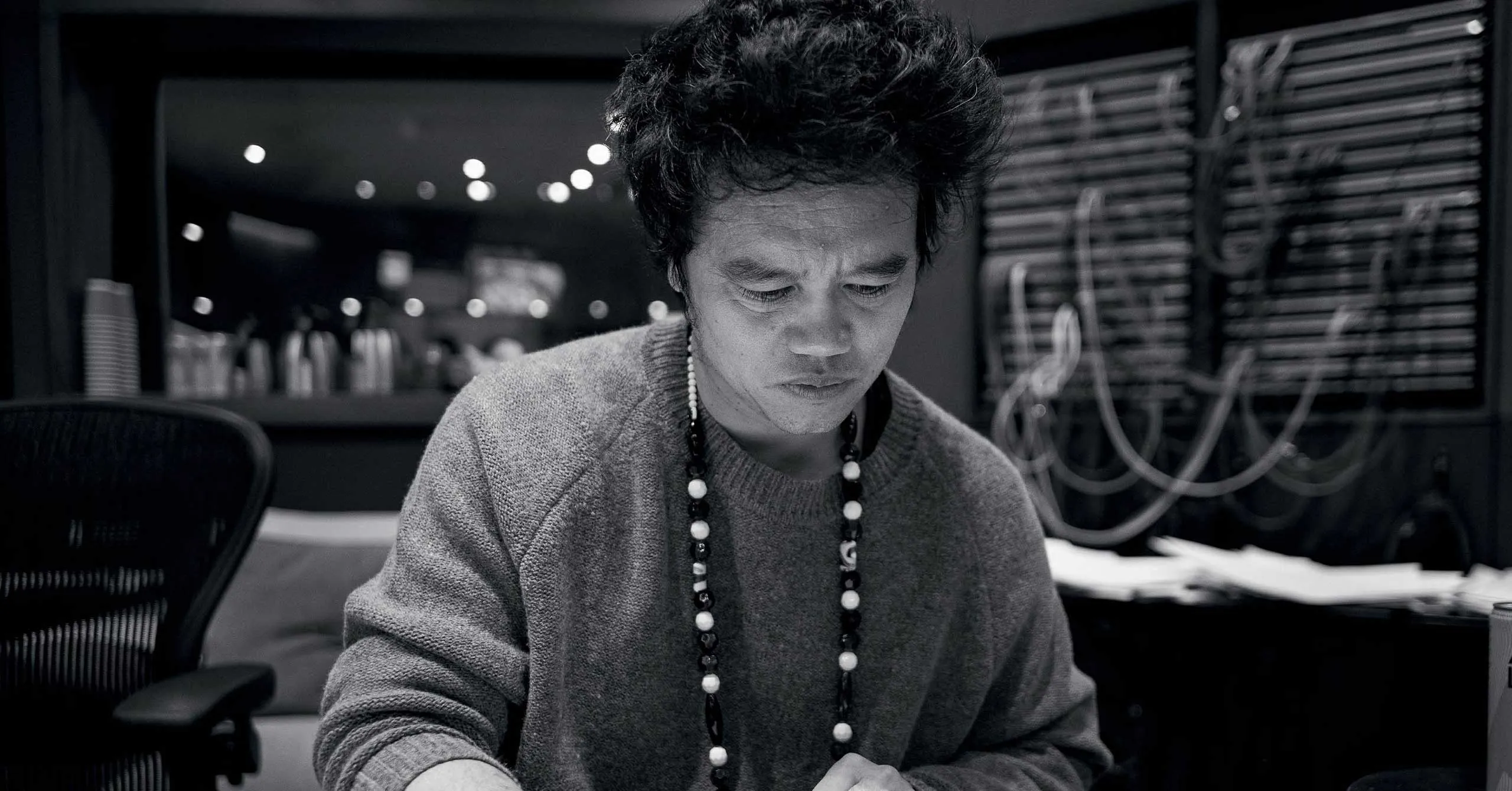 Person wearing a gray sweater and beaded necklace, focused on a task at a desk in a dim, equipment-filled studio.