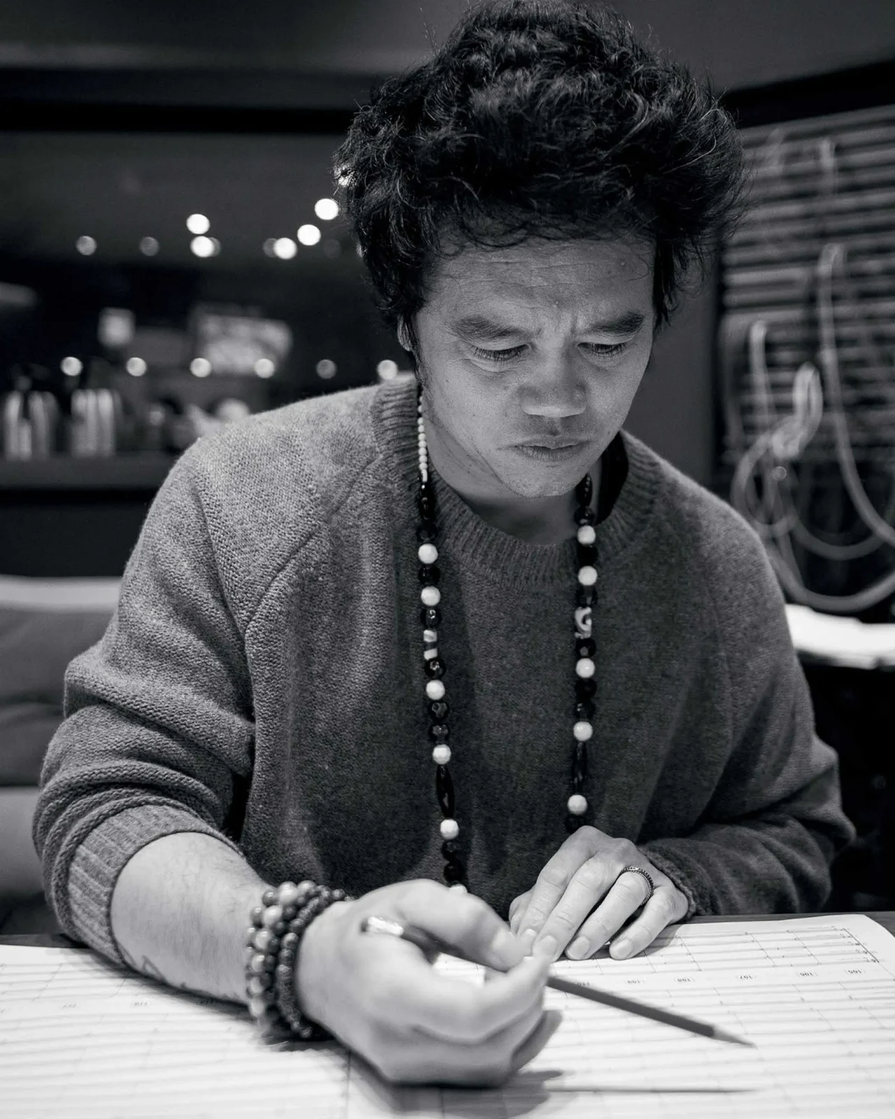 Person wearing a gray sweater and beaded necklace, focused on a task at a desk in a dim, equipment-filled studio.