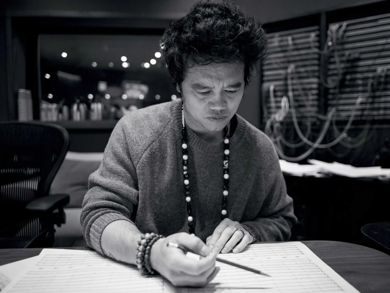 Person wearing a gray sweater and beaded necklace, focused on a task at a desk in a dim, equipment-filled studio.
