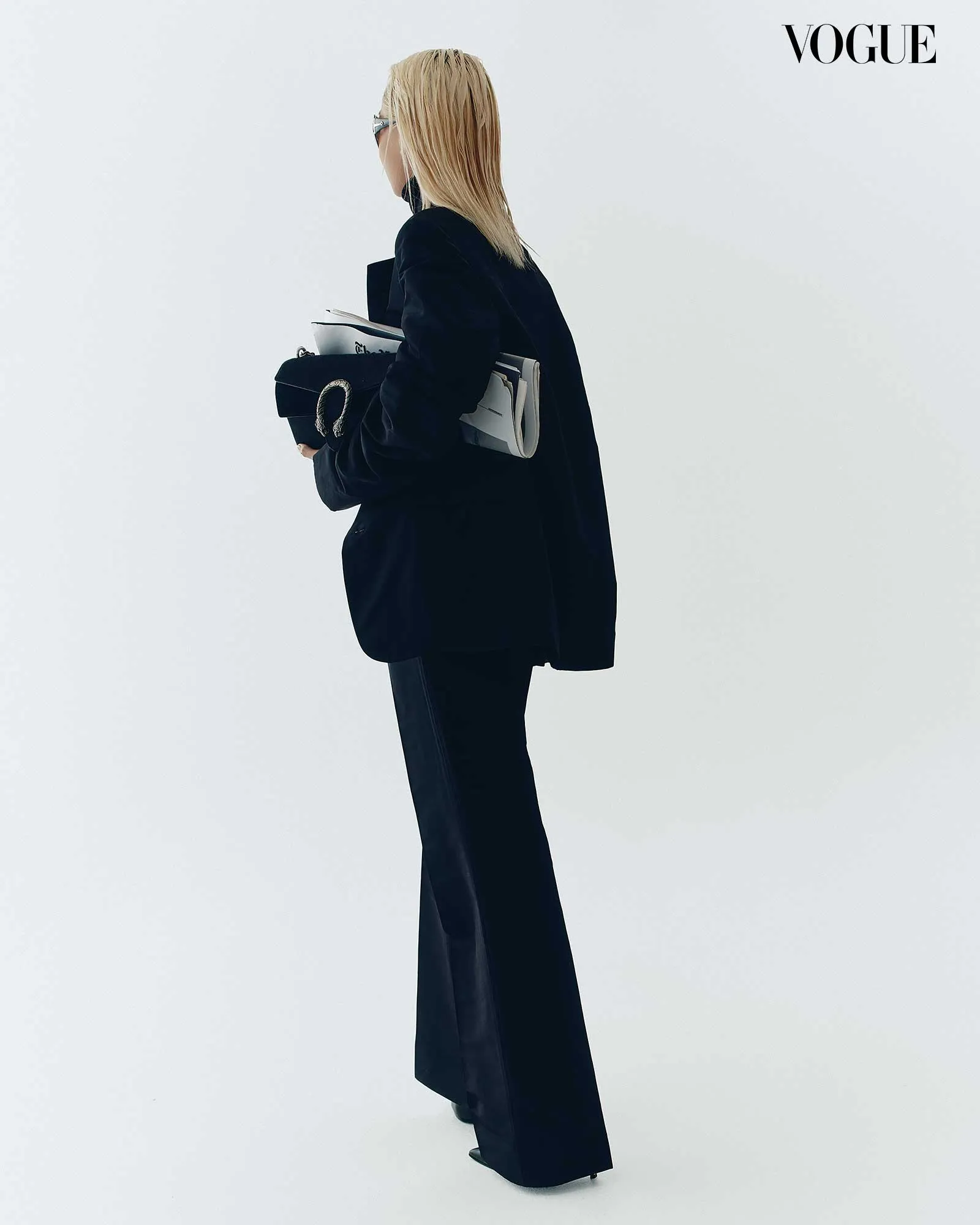Side profile of a woman in a black coat holding magazines and a handbag, with the Vogue logo in the top-right corner.