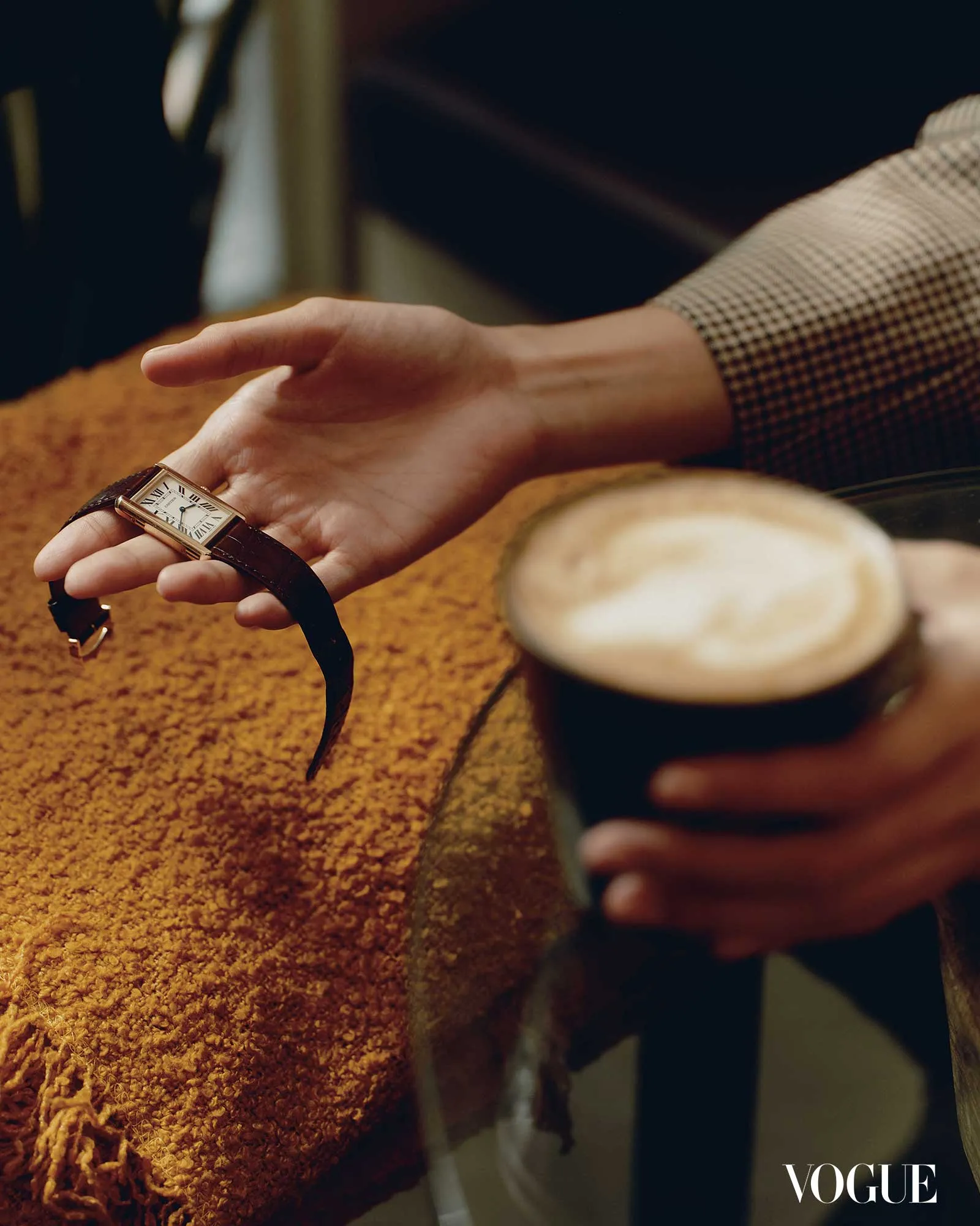 Close-up of a person’s hand with a rectangular gold watch resting on a textured brown scarf, a coffee cup in the foreground, Vogue logo in the lower right corner.