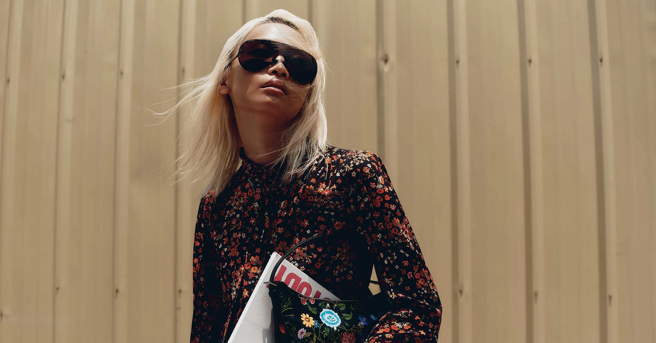 Woman with blonde hair wearing a black floral dress and oversized sunglasses, posing outdoors against a beige wall while holding papers/magazines