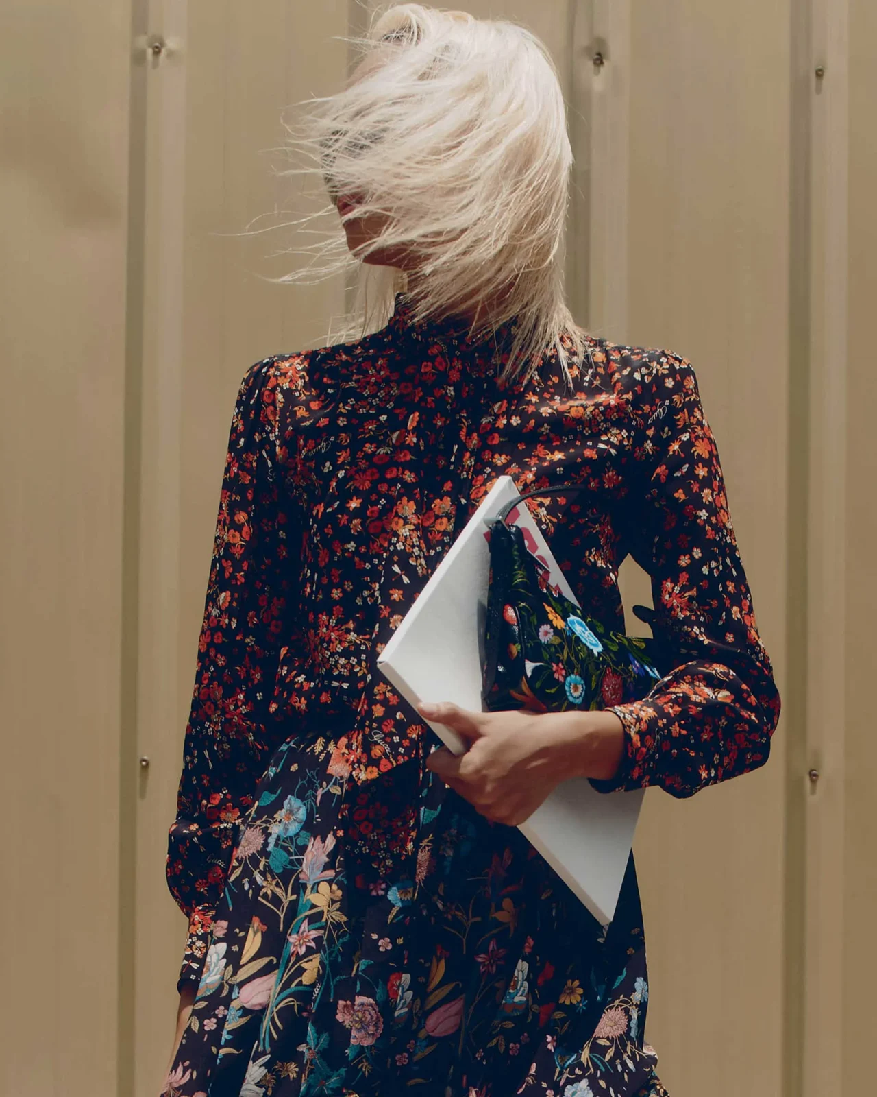 Woman with blonde hair wearing a black floral dress and oversized sunglasses, posing outdoors against a beige wall while holding papers/magazines