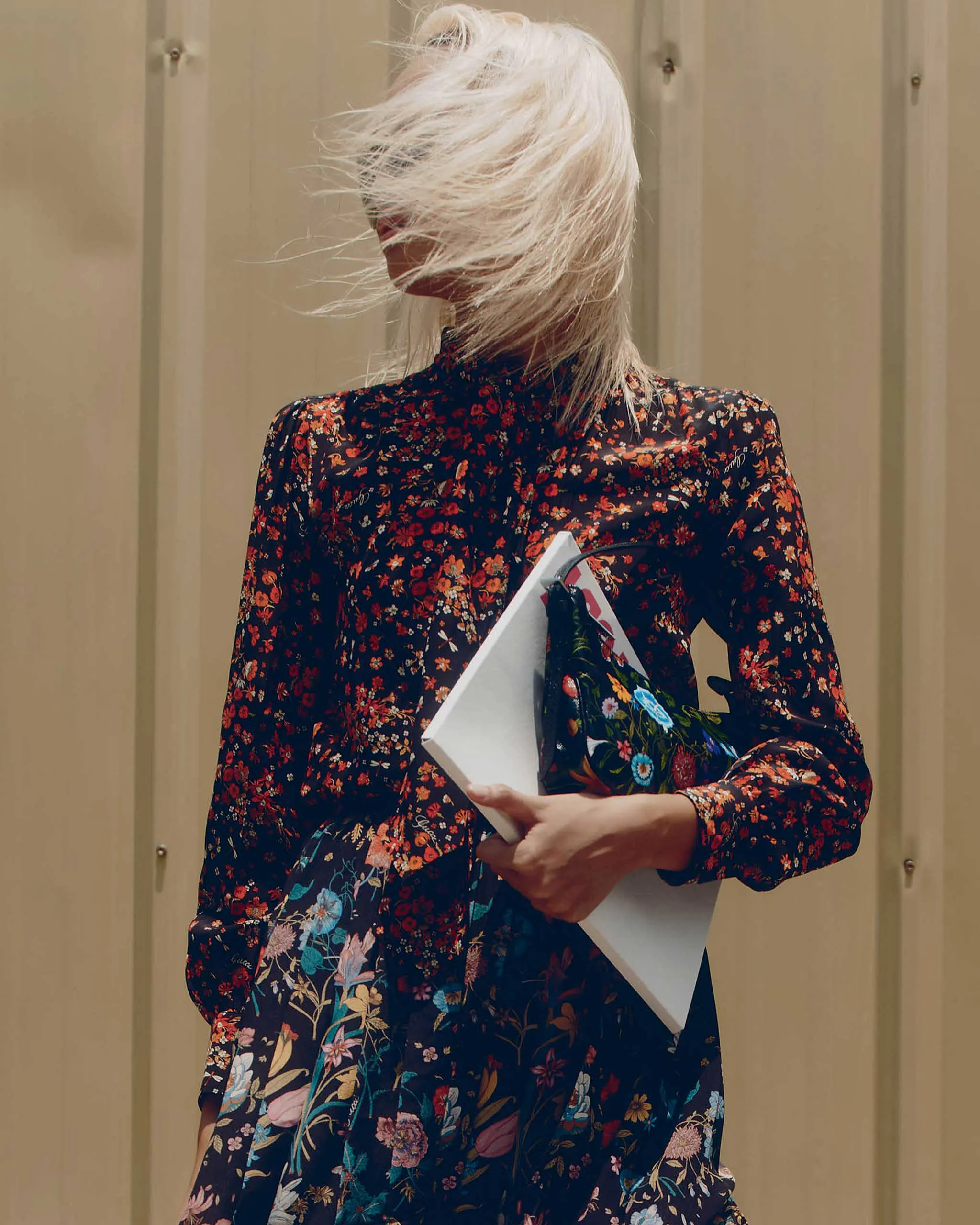 Person wearing a dark floral dress with wind-blown blonde hair, holding a white folder and a colorful bag in front of a beige wall.