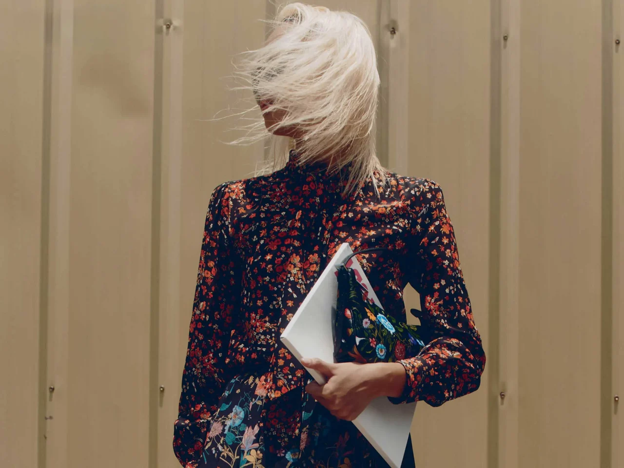 Woman with blonde hair wearing a black floral dress and oversized sunglasses, posing outdoors against a beige wall while holding papers/magazines