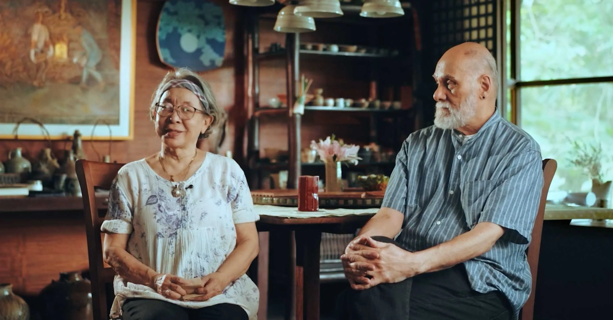 Elderly woman and man sit at a wooden dining table in a cozy home, chatting and smiling.