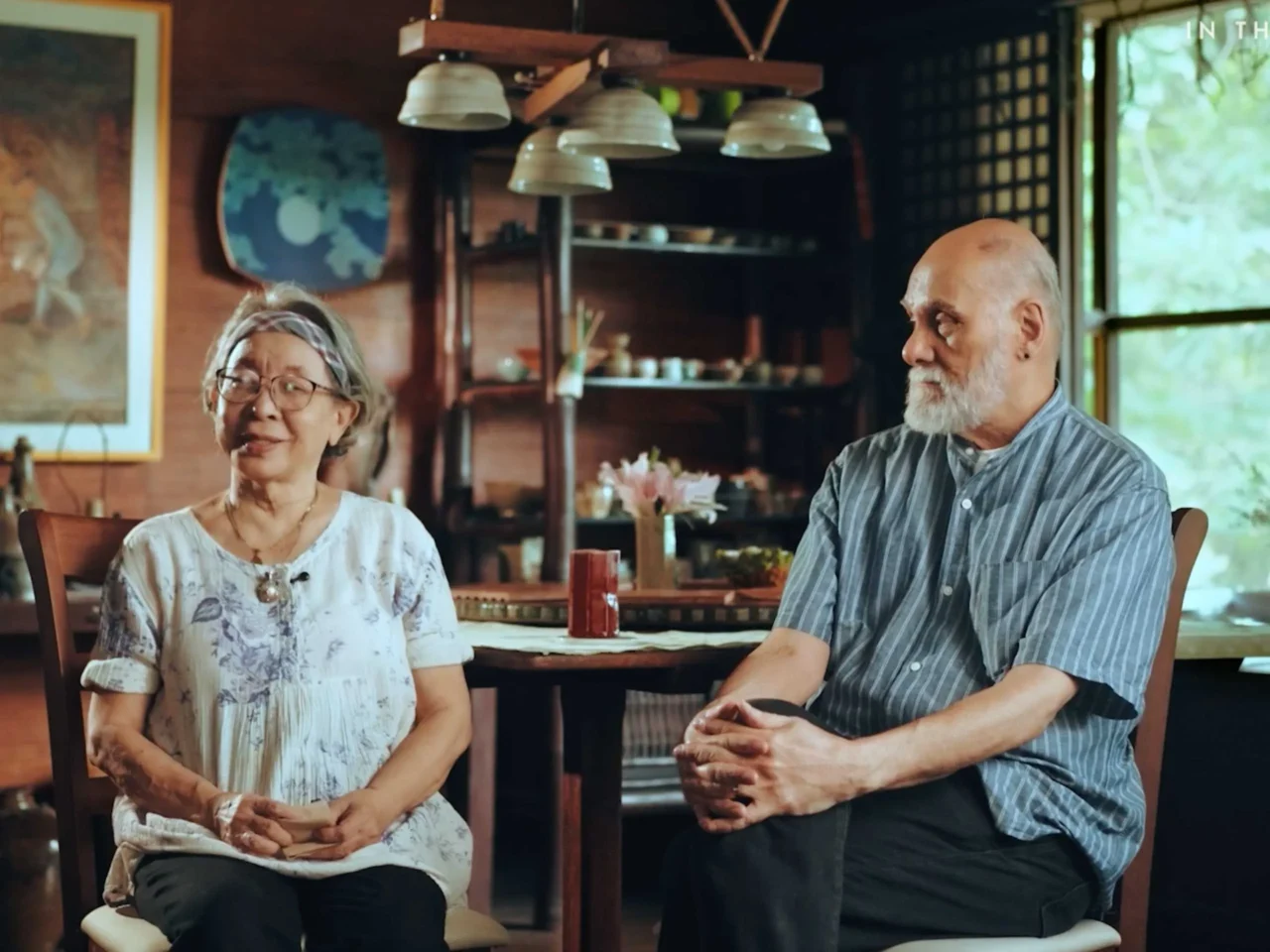 Elderly woman and man sit at a wooden dining table in a cozy home, chatting and smiling.