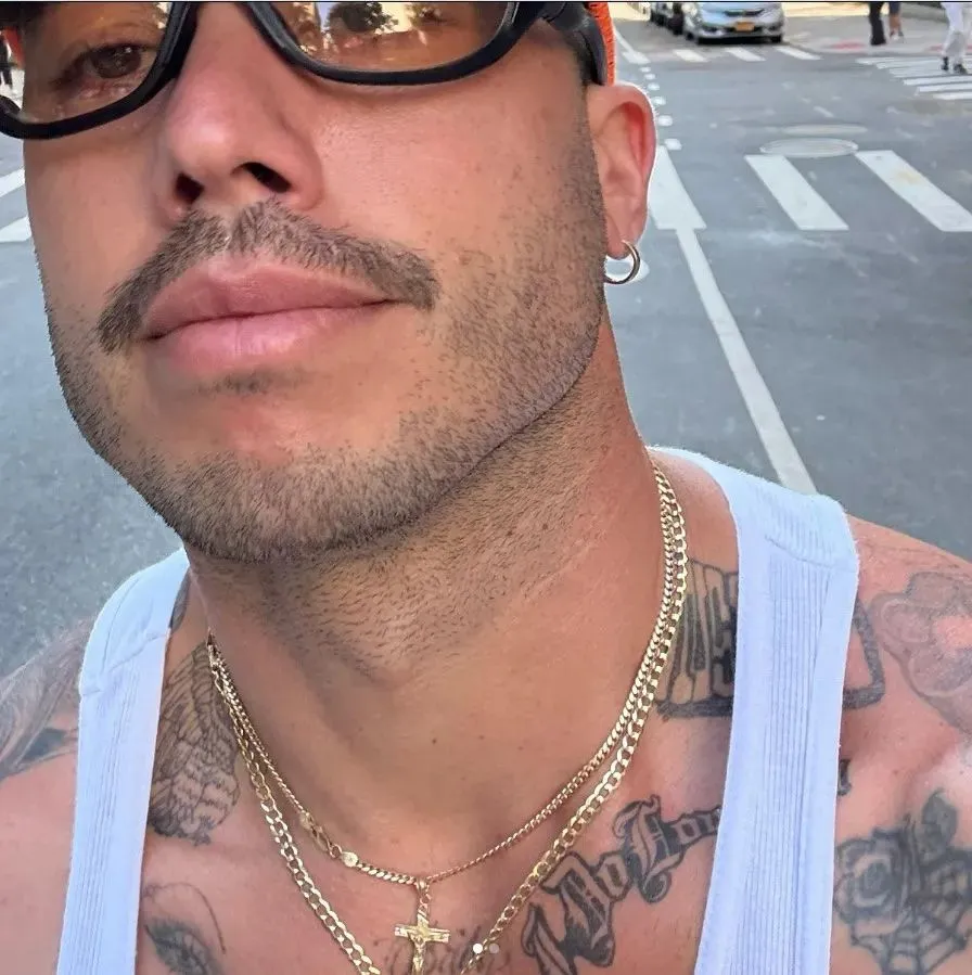Close-up of a tattooed man wearing sunglasses, a hoop earring, and gold chains on a white tank top, with a city street behind him.