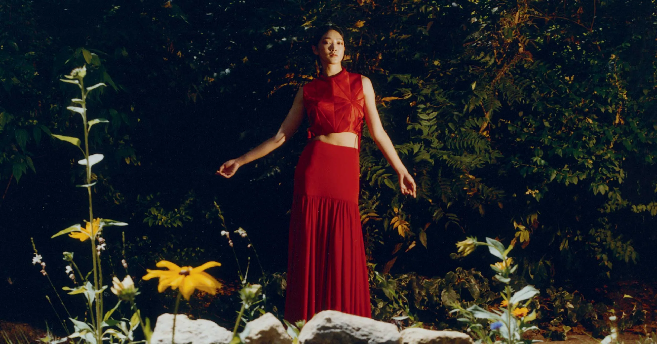 Woman in a red outfit standing in a sunlit garden among green foliage and yellow flowers.