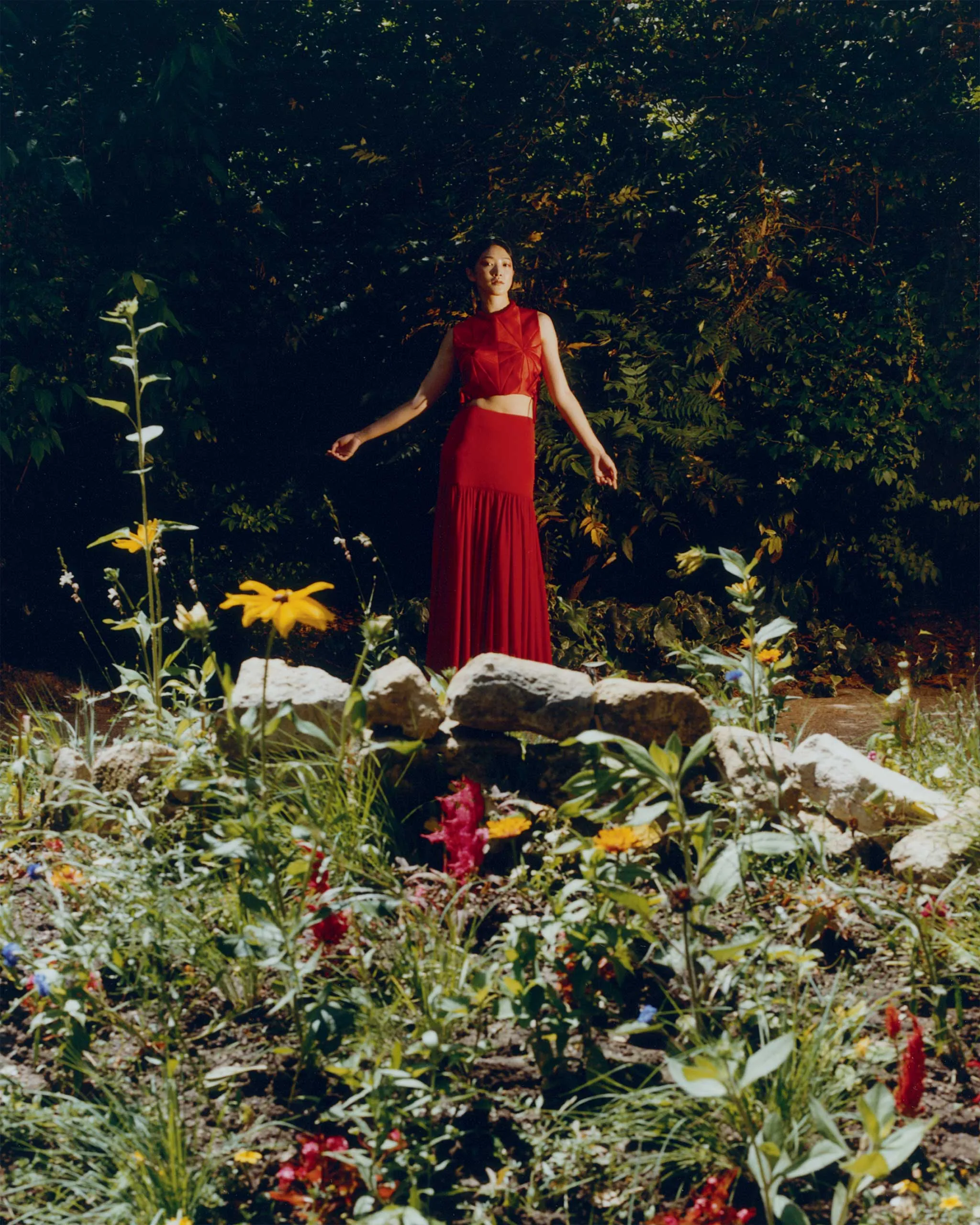 A person in a bright red sleeveless dress stands among tall green plants in a garden.