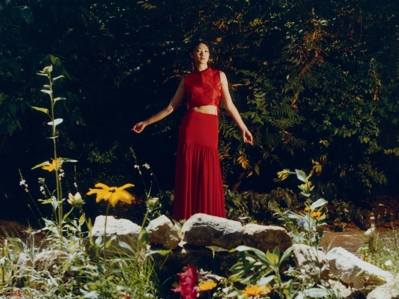 Woman in a red outfit standing in a sunlit garden among green foliage and yellow flowers.