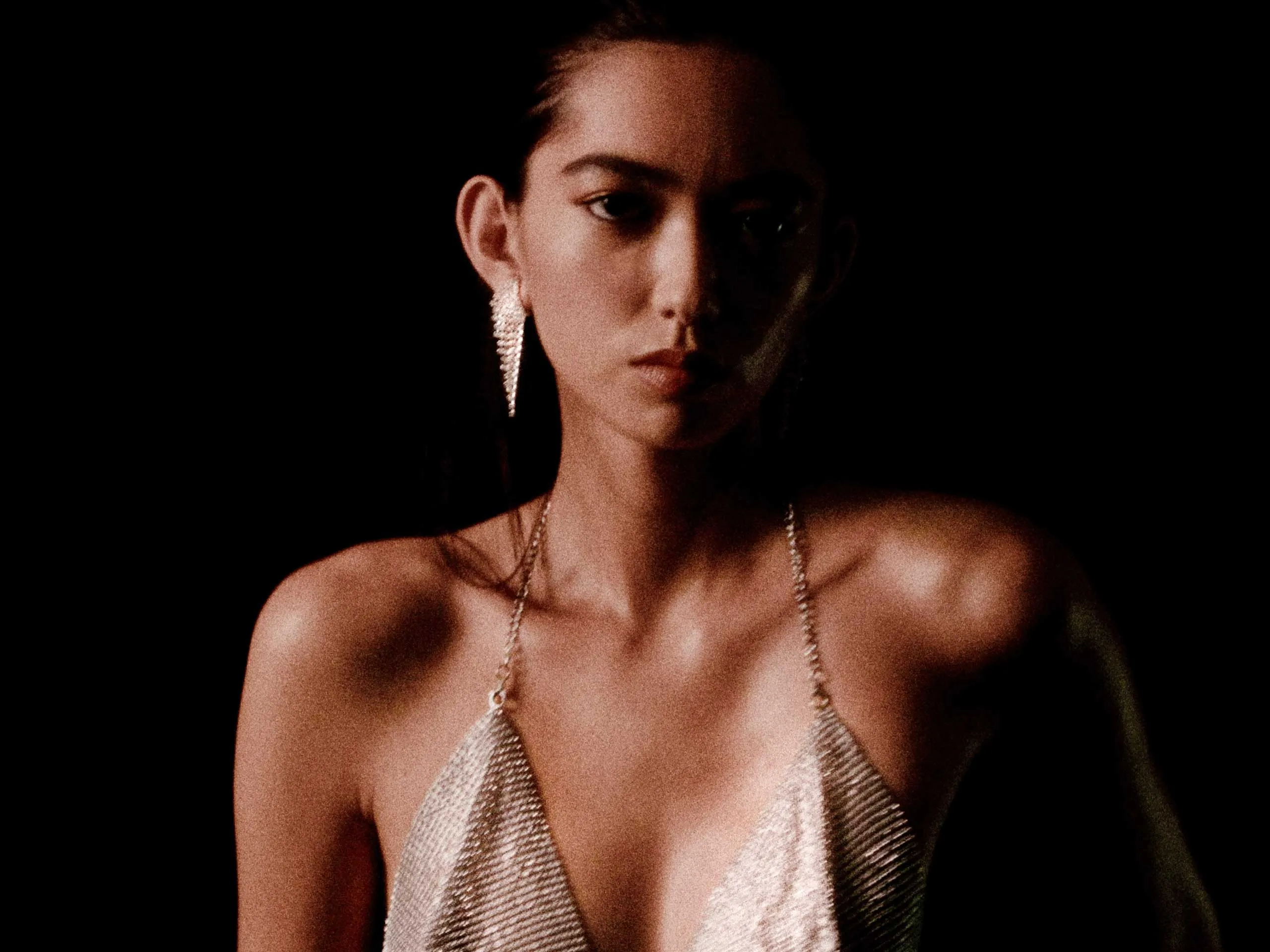Close-up of a person wearing a metallic silver halter top and long dangling earrings against a black background, low light.
