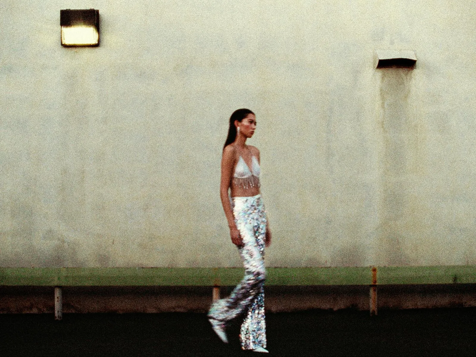 Woman in a white crop top and long floral skirt walks in profile along a beige concrete wall.
