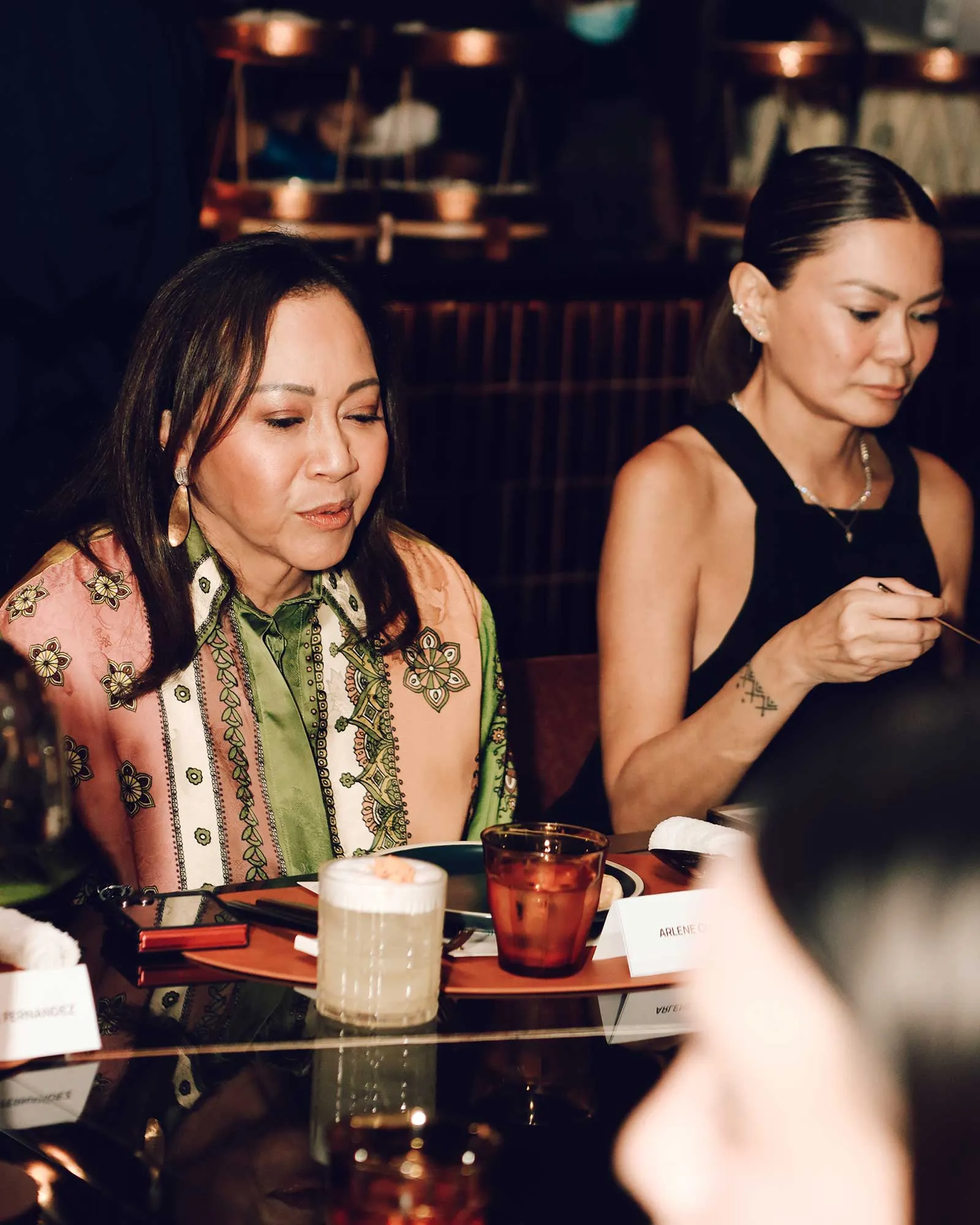 Two women sit at a dinner table in a dimly lit restaurant, engaged in conversation with plates and drinks in front of them.
