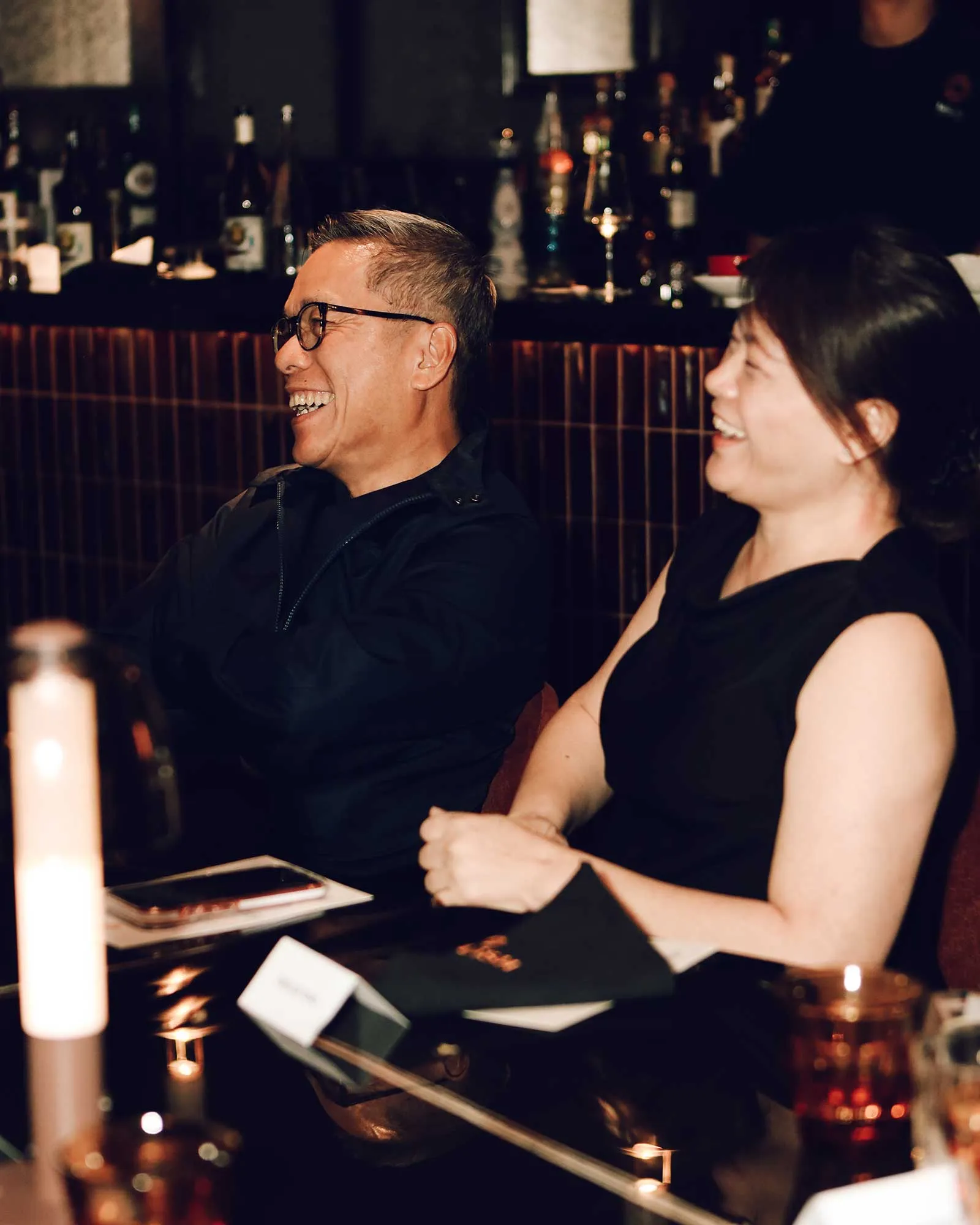 Two friends seated at a candlelit bar/table, laughing together as they share a moment.