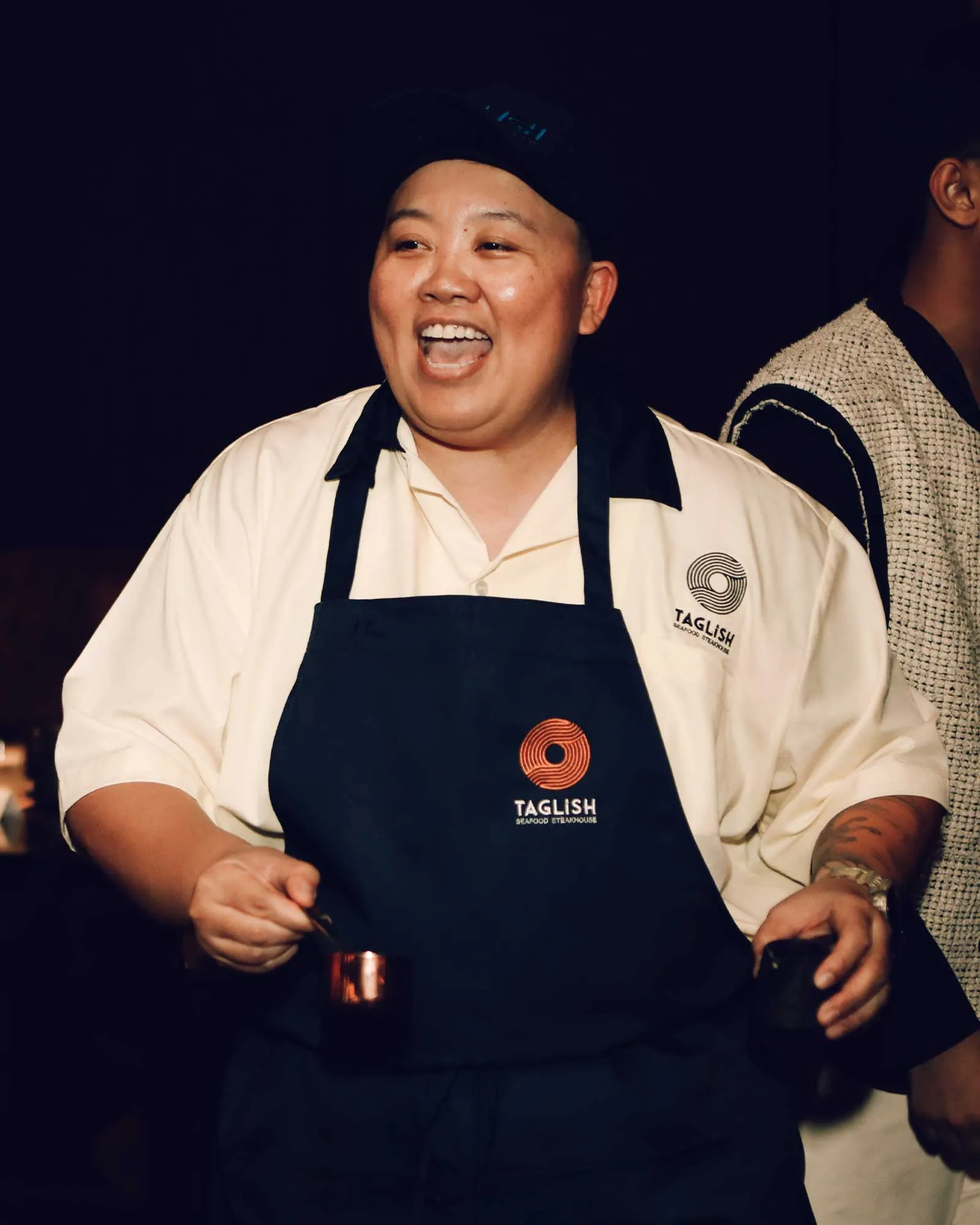 Chef in a white shirt and black apron laughs with mouth open, Taglish logo visible on the apron.