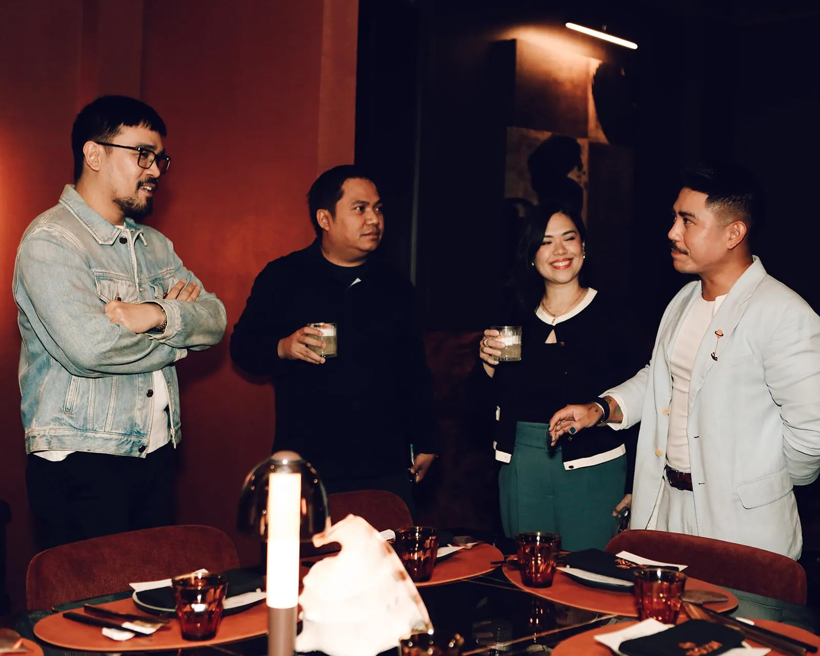 Four friends stand and chat in a dimly lit restaurant, each holding drinks and smiling at one another.