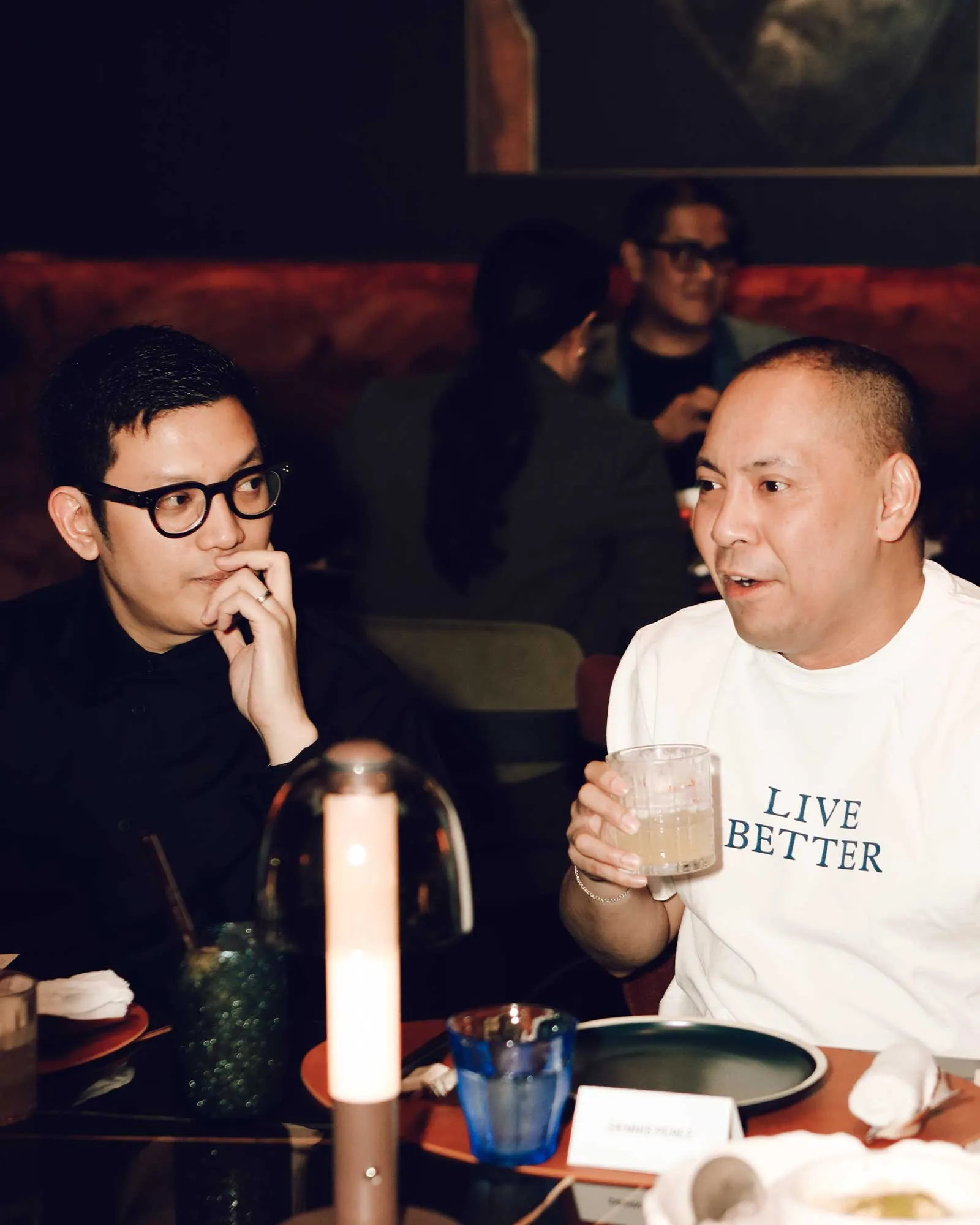 Two men converse at a dim restaurant table; left wears glasses, right in a white shirt, candle glowing between them.