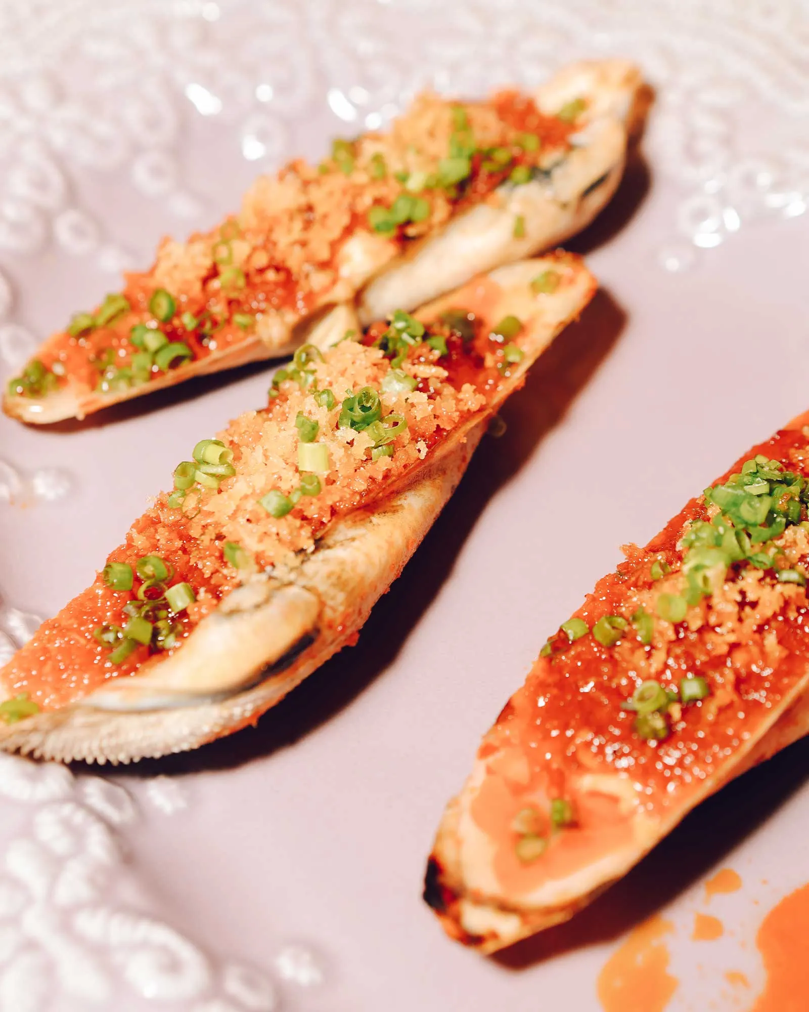 Open-faced crostini topped with a red spicy sauce, crunchy breadcrumbs, and chopped green onions on a pale plate.